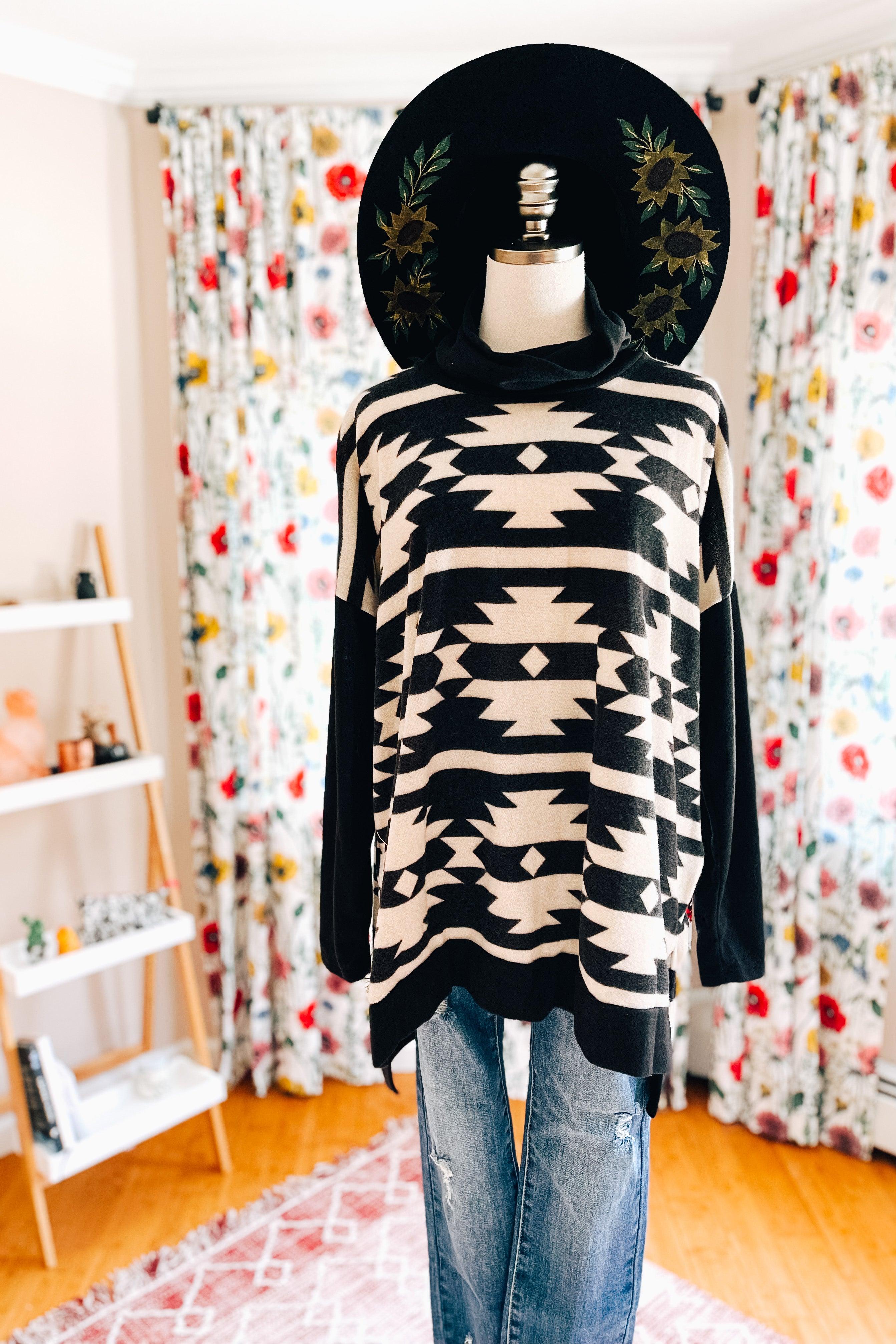 Mannequin wearing black and white geometric patterned cowl neck pullover with blue distressed jeans and wide black hat indoors