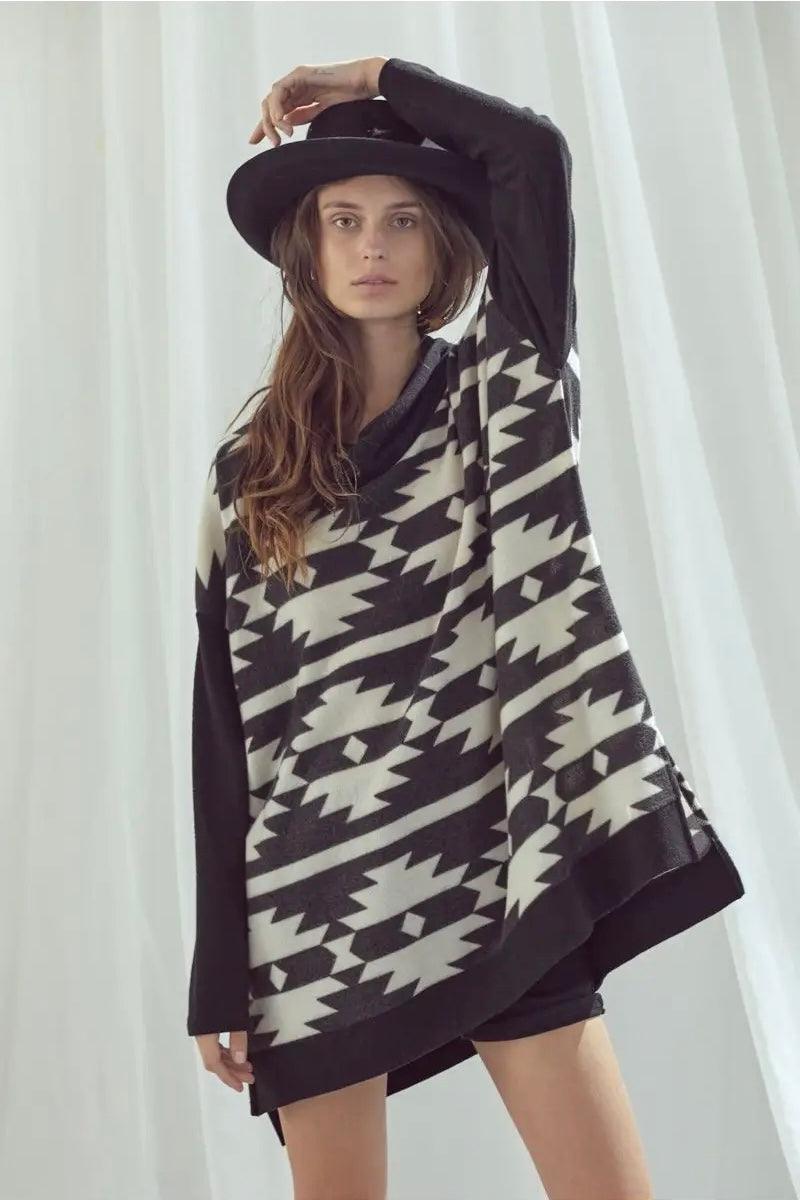 Model wearing oversized black and white geometric patterned cowl neck pullover with wide-brim black hat indoors