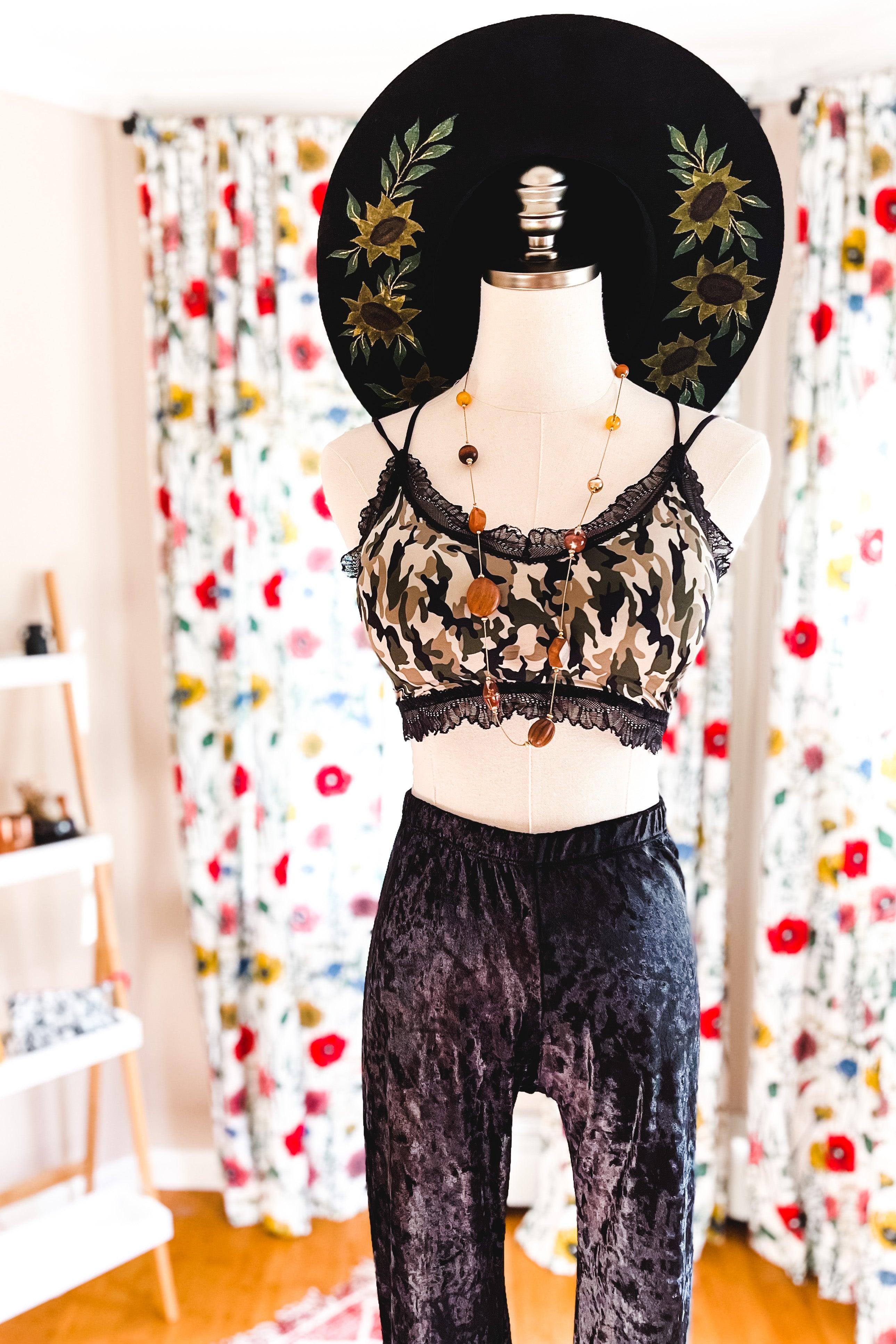 Mannequin wearing camo bralette with black lace trim paired with black crushed velvet pants and floral wide-brim hat