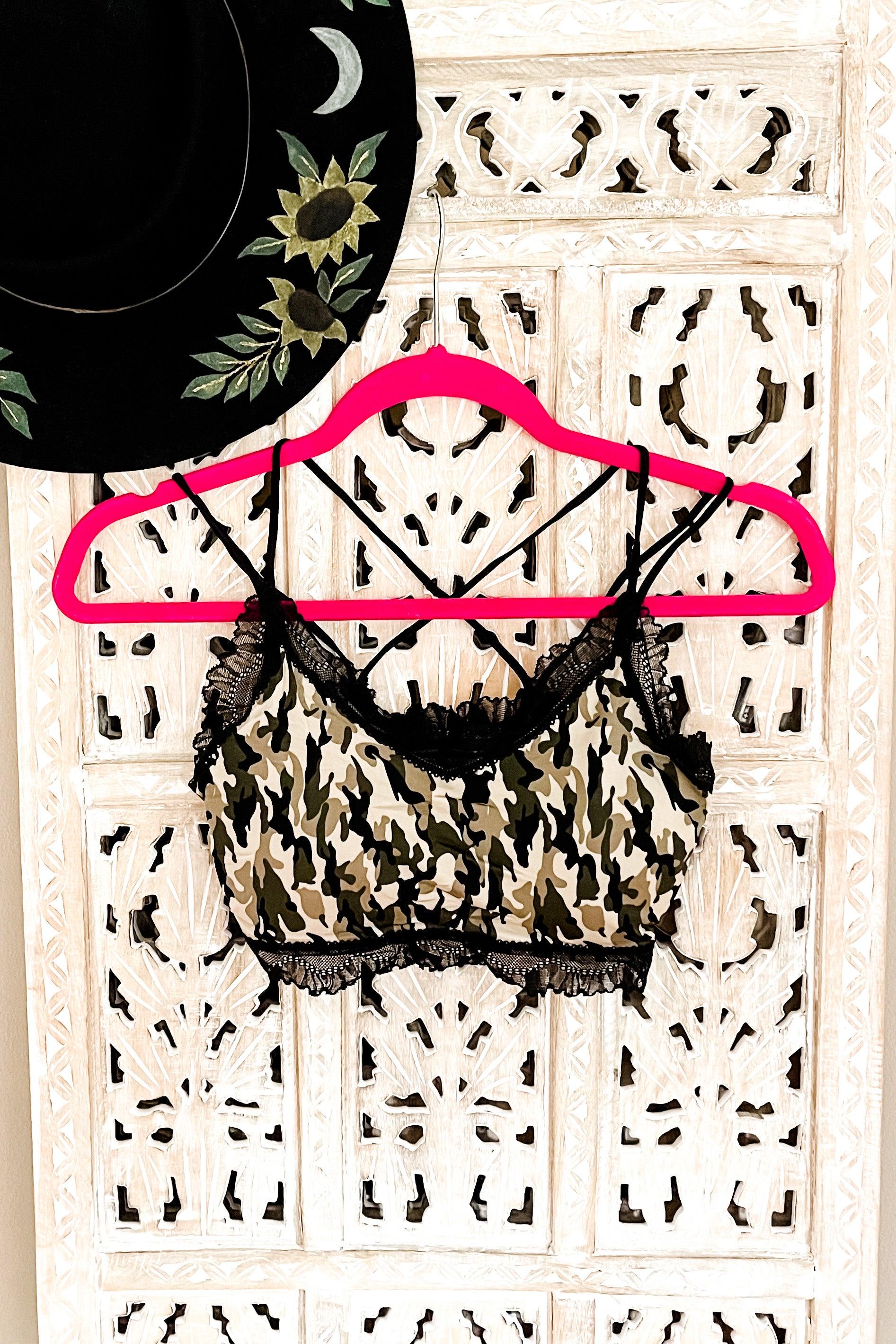 Camo bralette with black lace trim hanging on bright pink hanger beside black hat with sunflower embroidery on white carved wood panel