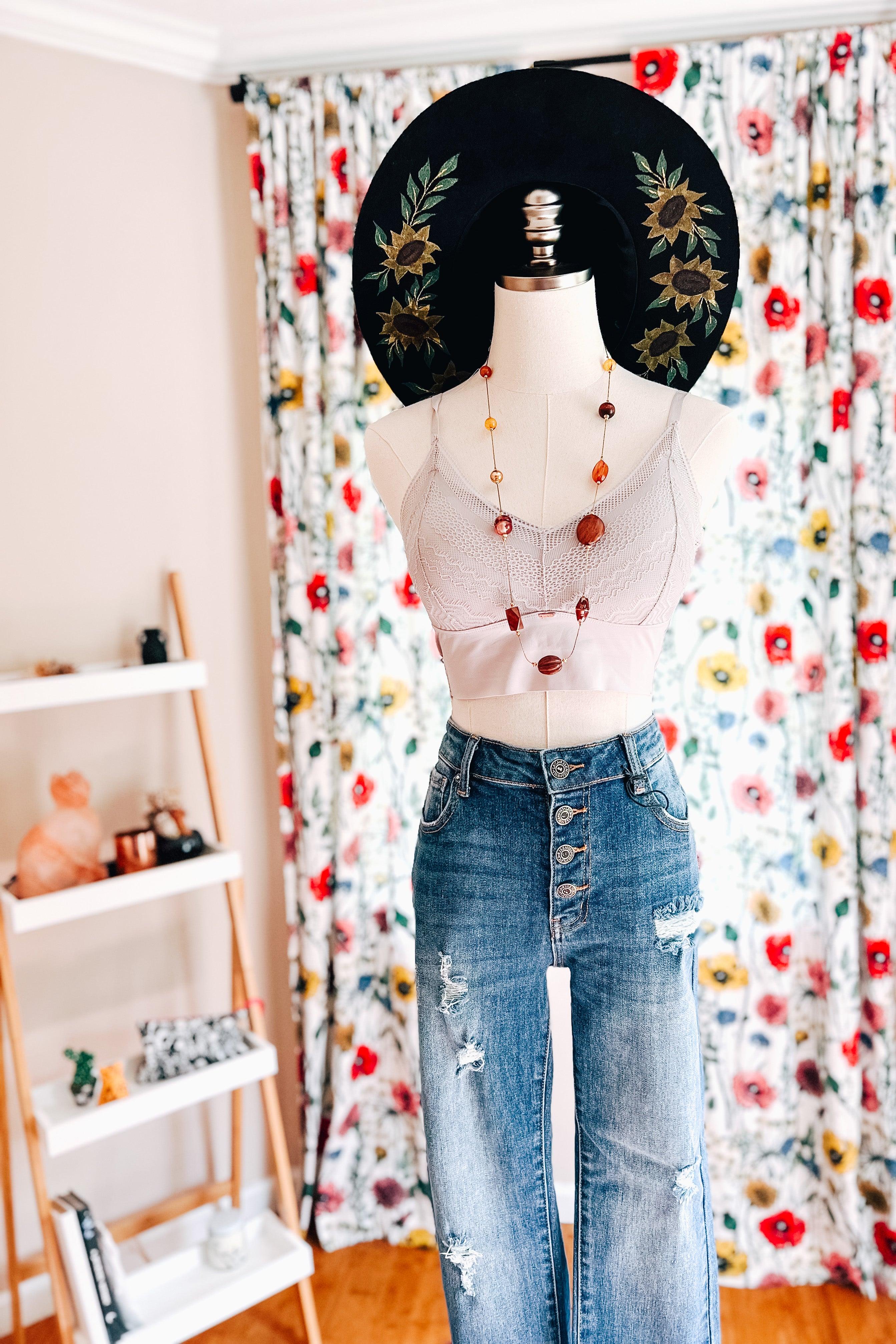 Mannequin wearing silver bralette and high-waisted distressed jeans with black wide-brim hat and beaded necklace indoors