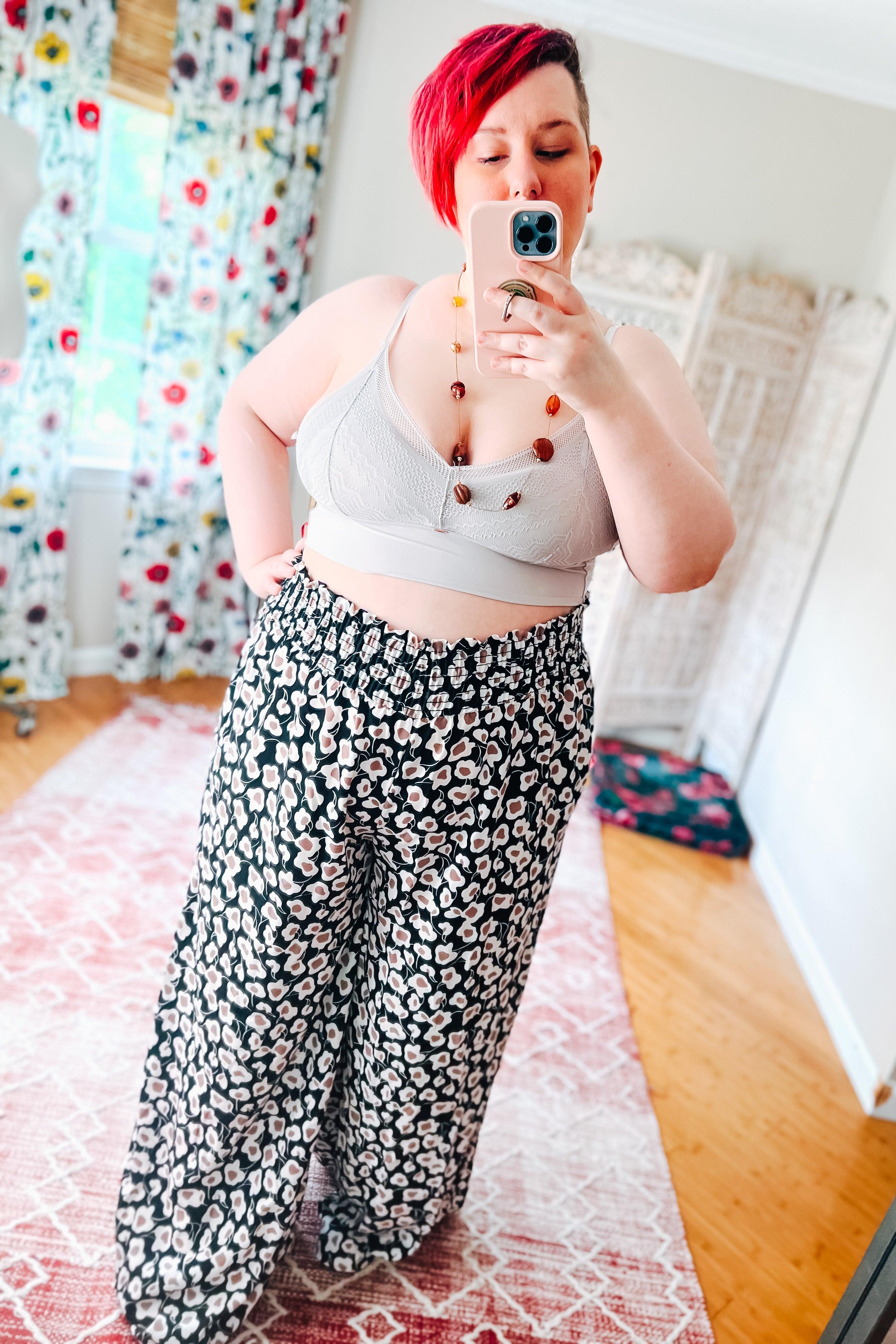 Plus size woman with red hair wearing silver textured bralette and black floral wide-leg pants taking a selfie in mirror