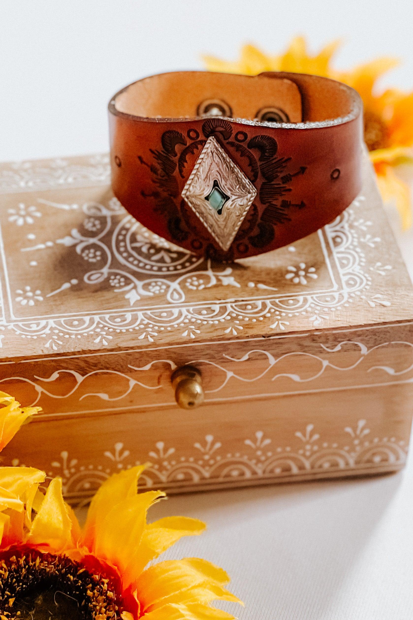 Leather cuff bracelet with engraved diamond-shaped silver centerpiece displayed on carved wooden box with sunflowers nearby