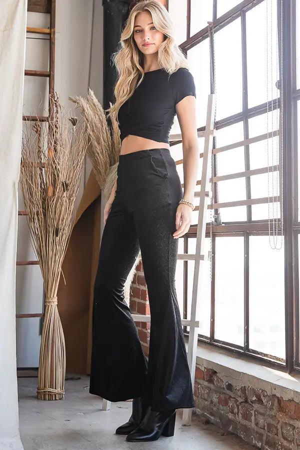 Blonde woman wearing black glittery bell bottoms and a fitted black crop top standing by large window in loft space