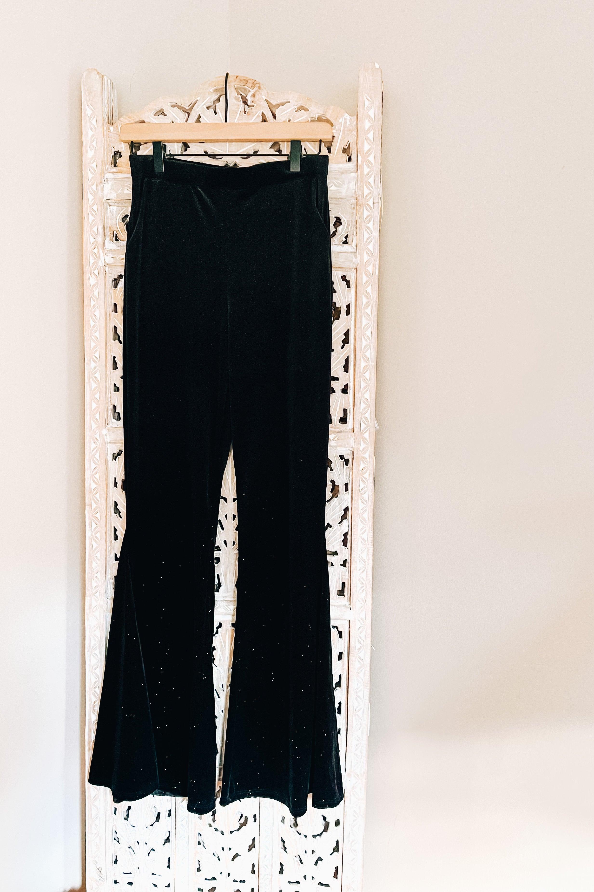 Black velvet bell bottoms with subtle glitter detail hanging on wooden hanger against decorative white screen