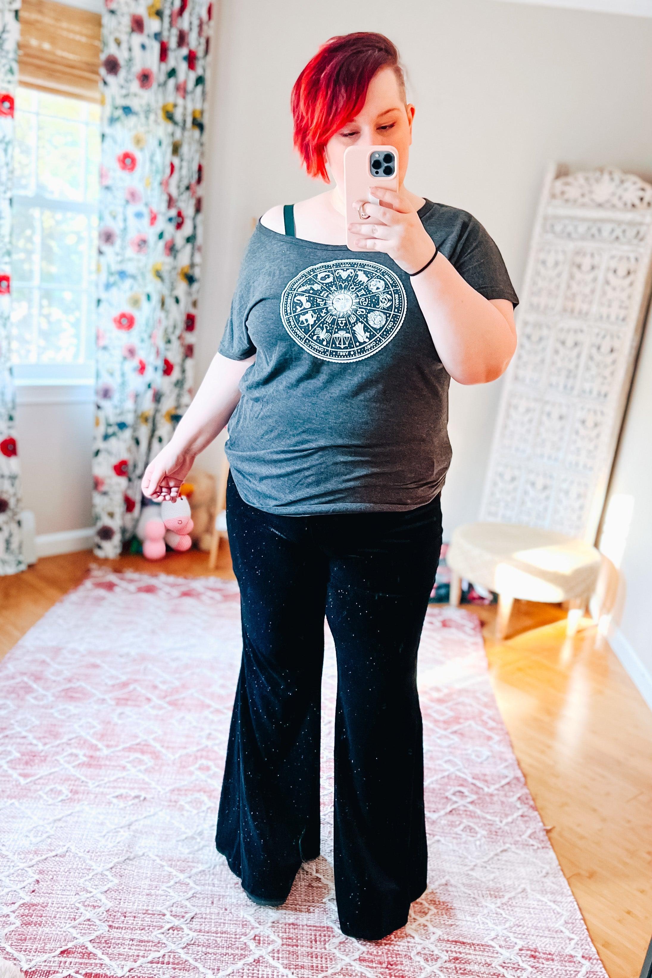 Woman with short red hair wearing black glitter disco bell bottoms and gray zodiac graphic t-shirt taking mirror selfie indoors