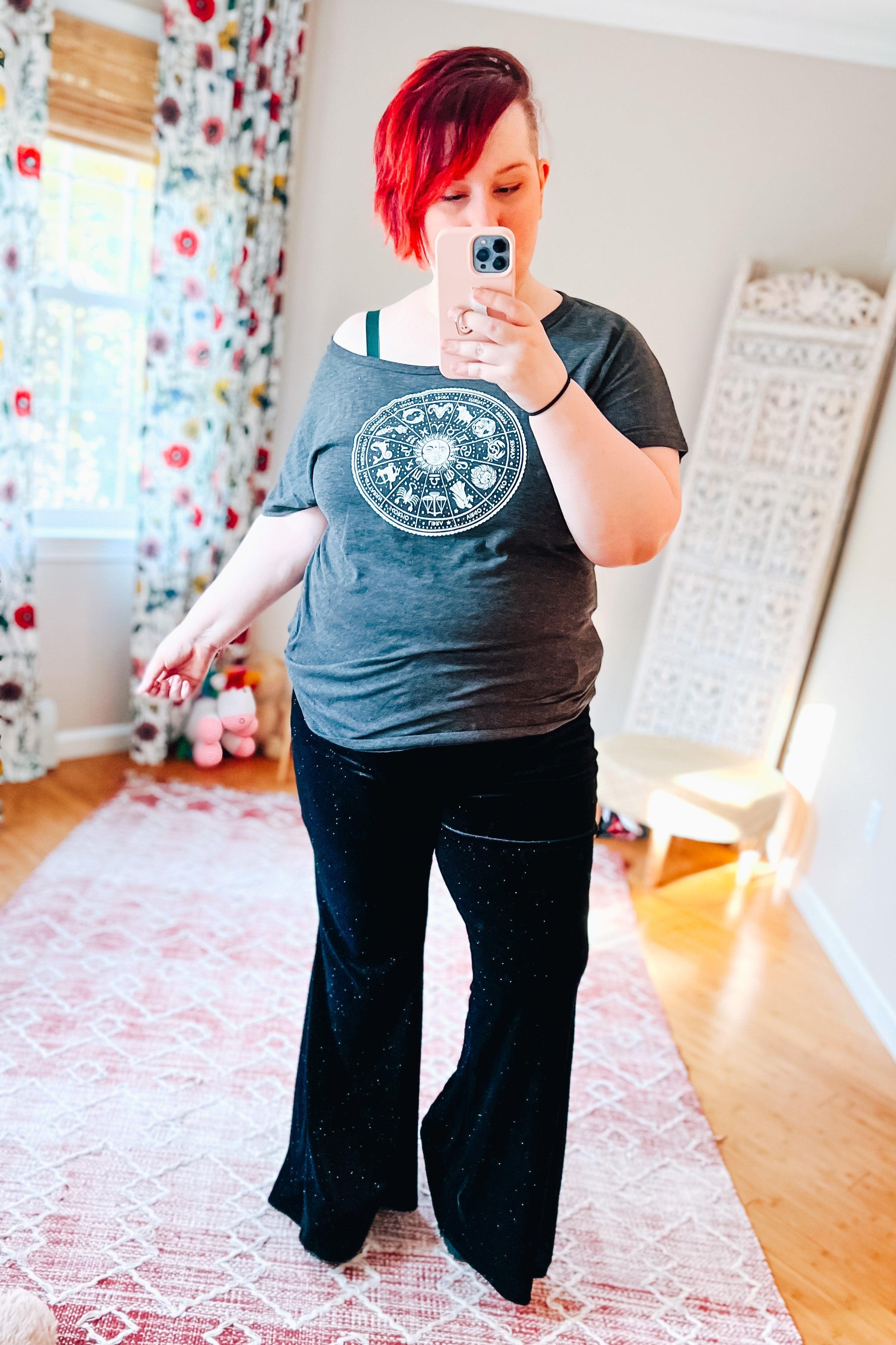 Plus size woman with short red hair wearing black glitter flare pants and gray zodiac graphic t-shirt taking mirror selfie indoors
