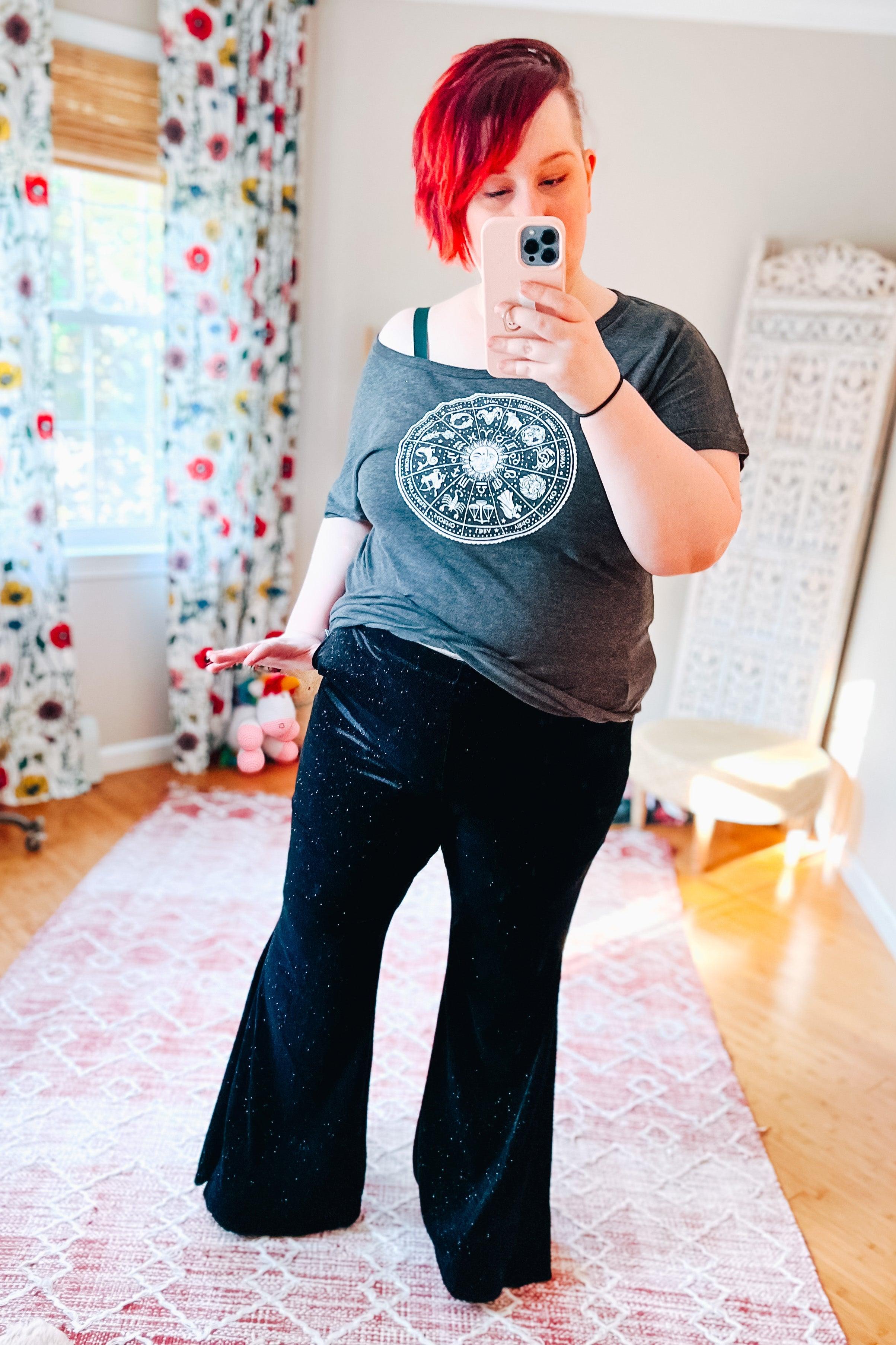 Plus size woman with short red hair wearing black glitter bell bottoms and gray zodiac graphic t-shirt taking mirror selfie indoors