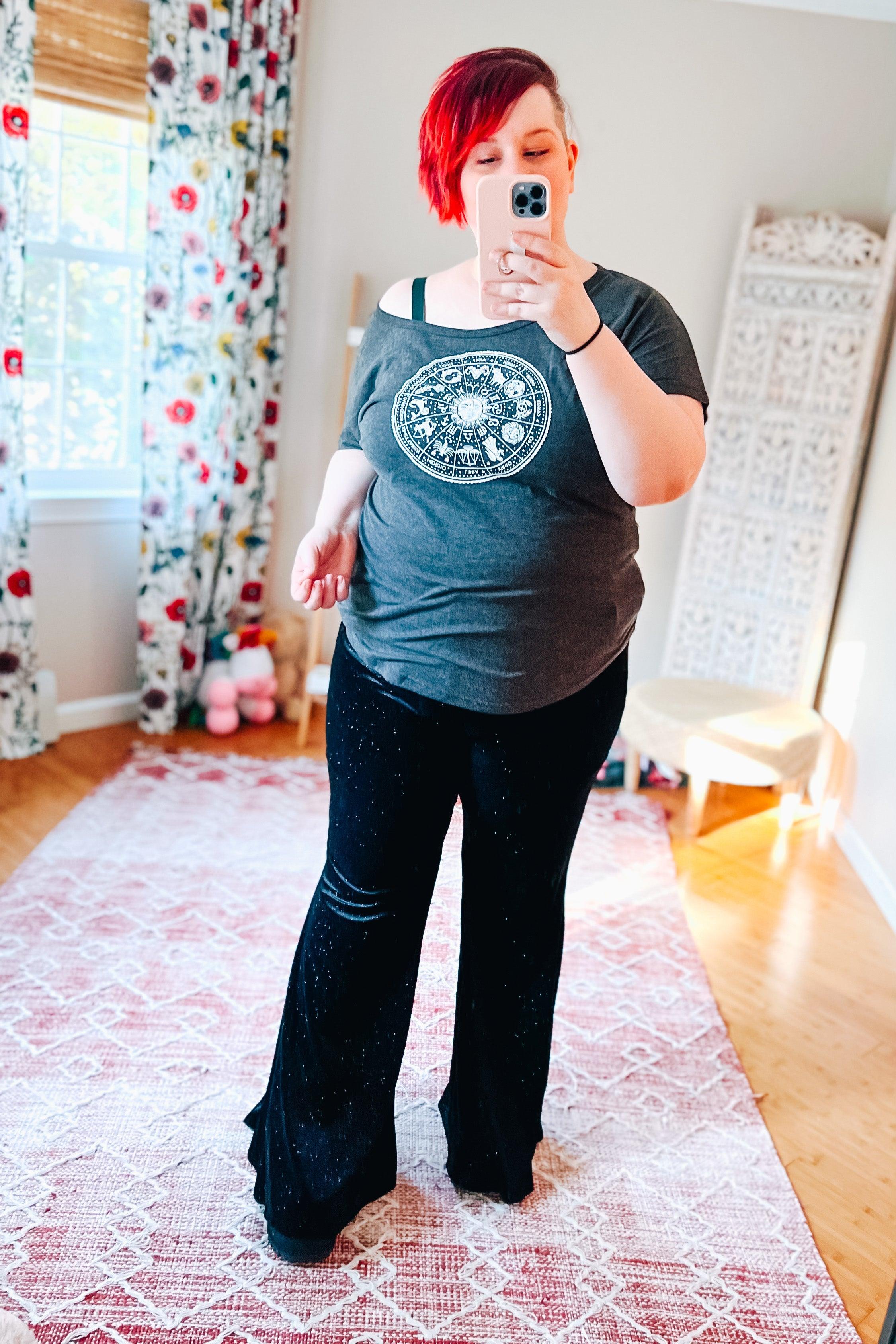 Plus size woman with short red hair wearing black glitter bell bottoms and gray graphic off-shoulder top taking mirror selfie indoors