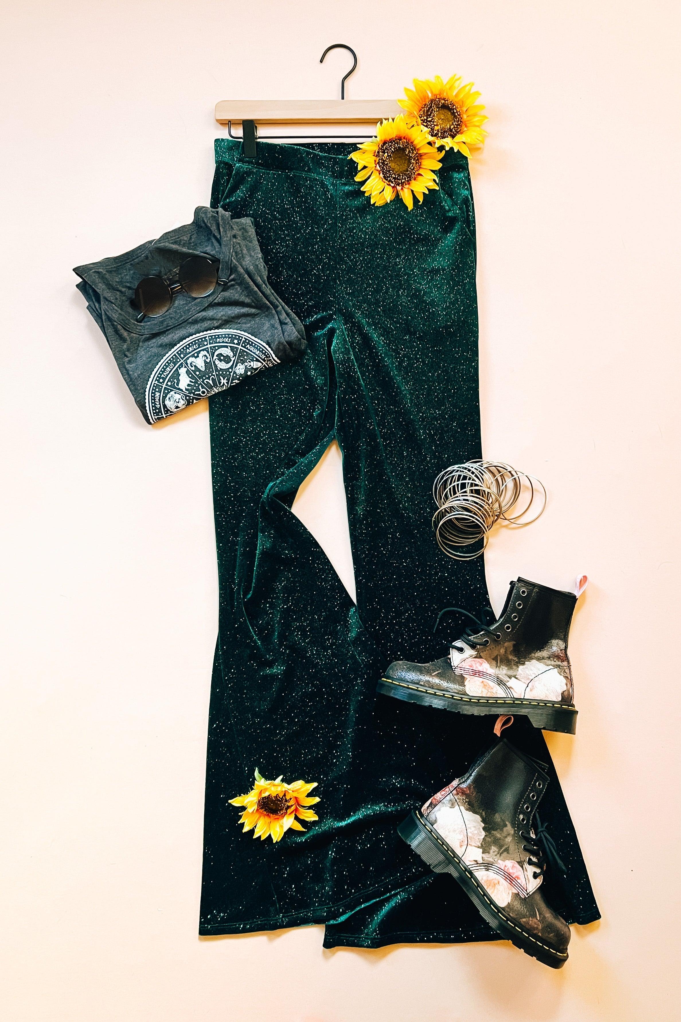 Evergreen glitter velvet bell bottoms styled with black graphic tee, floral combat boots, silver bracelets, and sunflowers on pale background