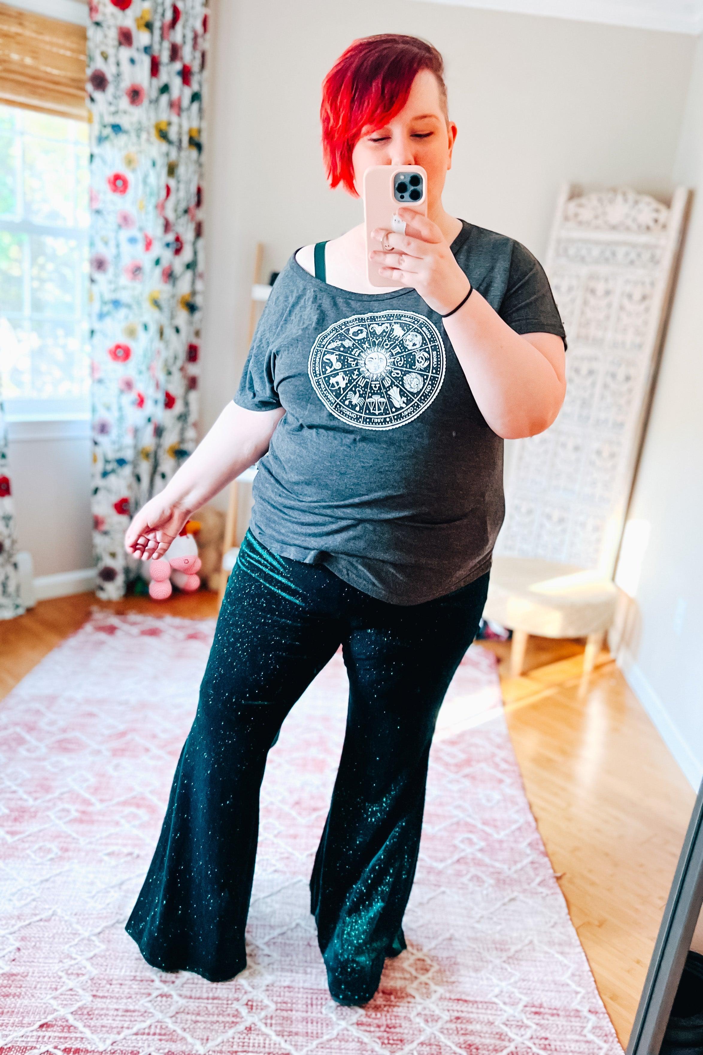 Woman taking mirror selfie wearing black glitter bell bottoms and dark grey zodiac print t-shirt in bright room