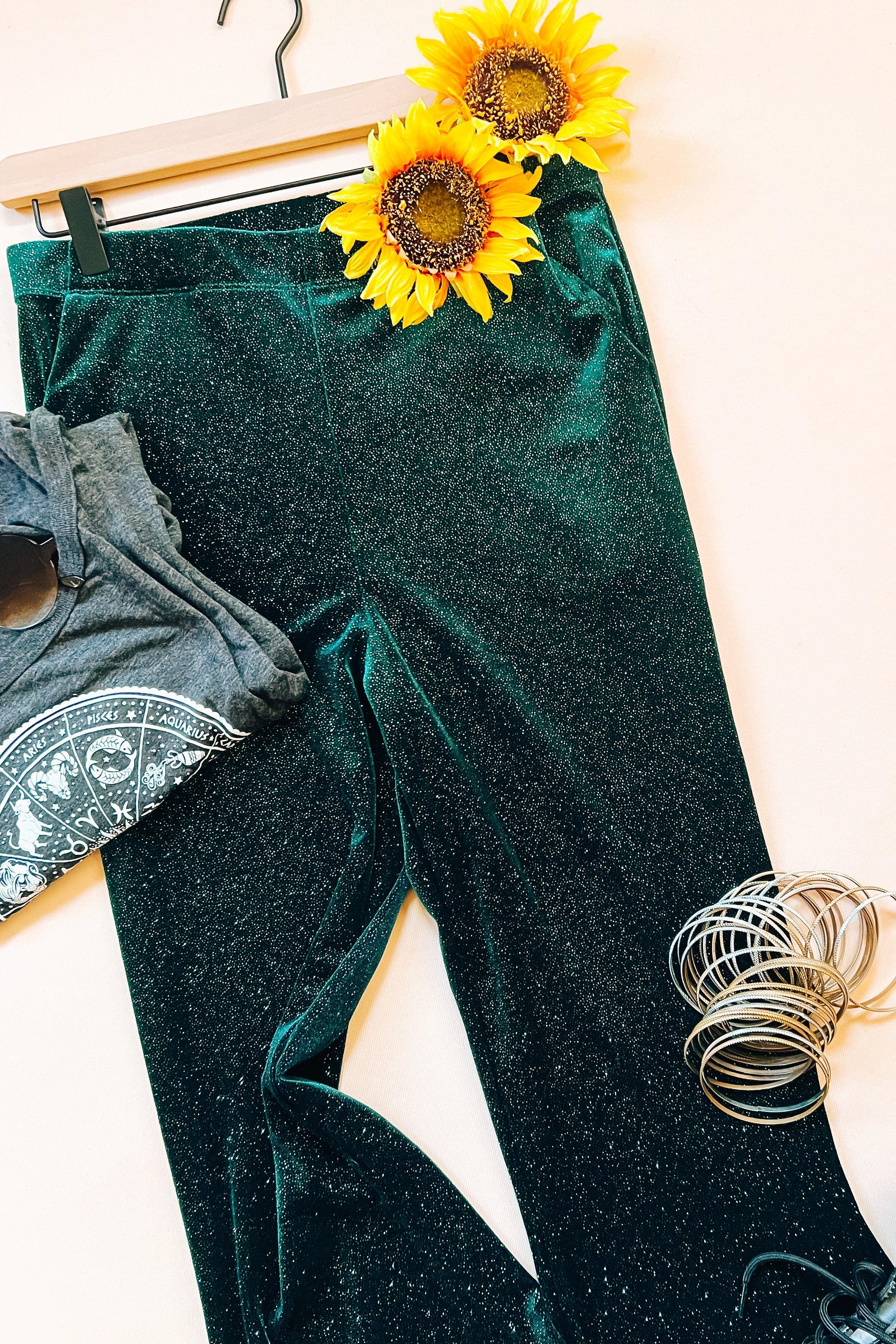Evergreen glitter velvet bell bottoms laid flat with grey zodiac tee, sunglasses, bangles, and sunflower decor