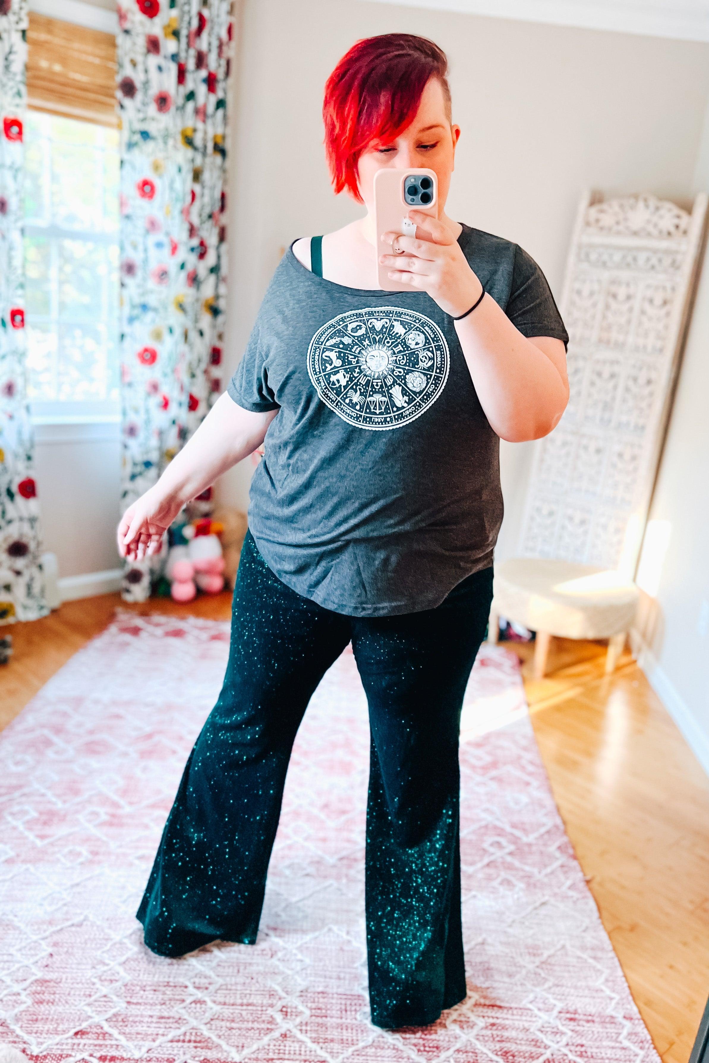 Woman wearing shimmering evergreen glitter bell bottoms paired with grey zodiac graphic tee taking a selfie indoors