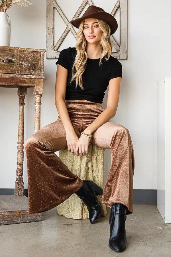 Woman seated wearing gold glitter velvet bell bottoms paired with black cropped top and brown hat indoors