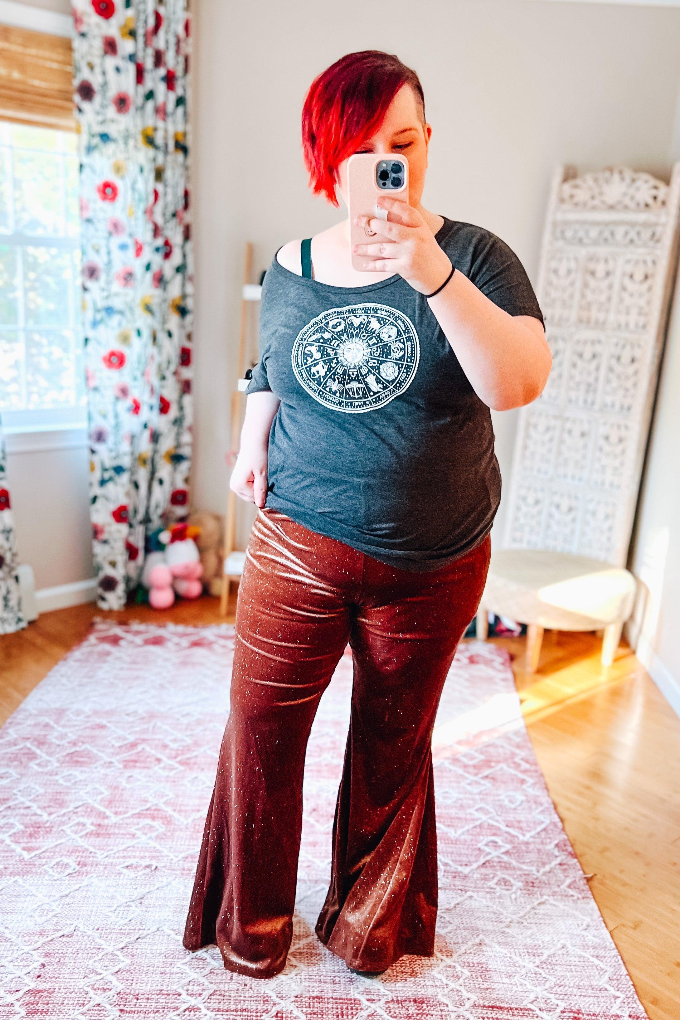 Woman wearing rust velvet glitter bell bottoms and dark grey zodiac graphic tee taking selfie in bright room with floral curtains