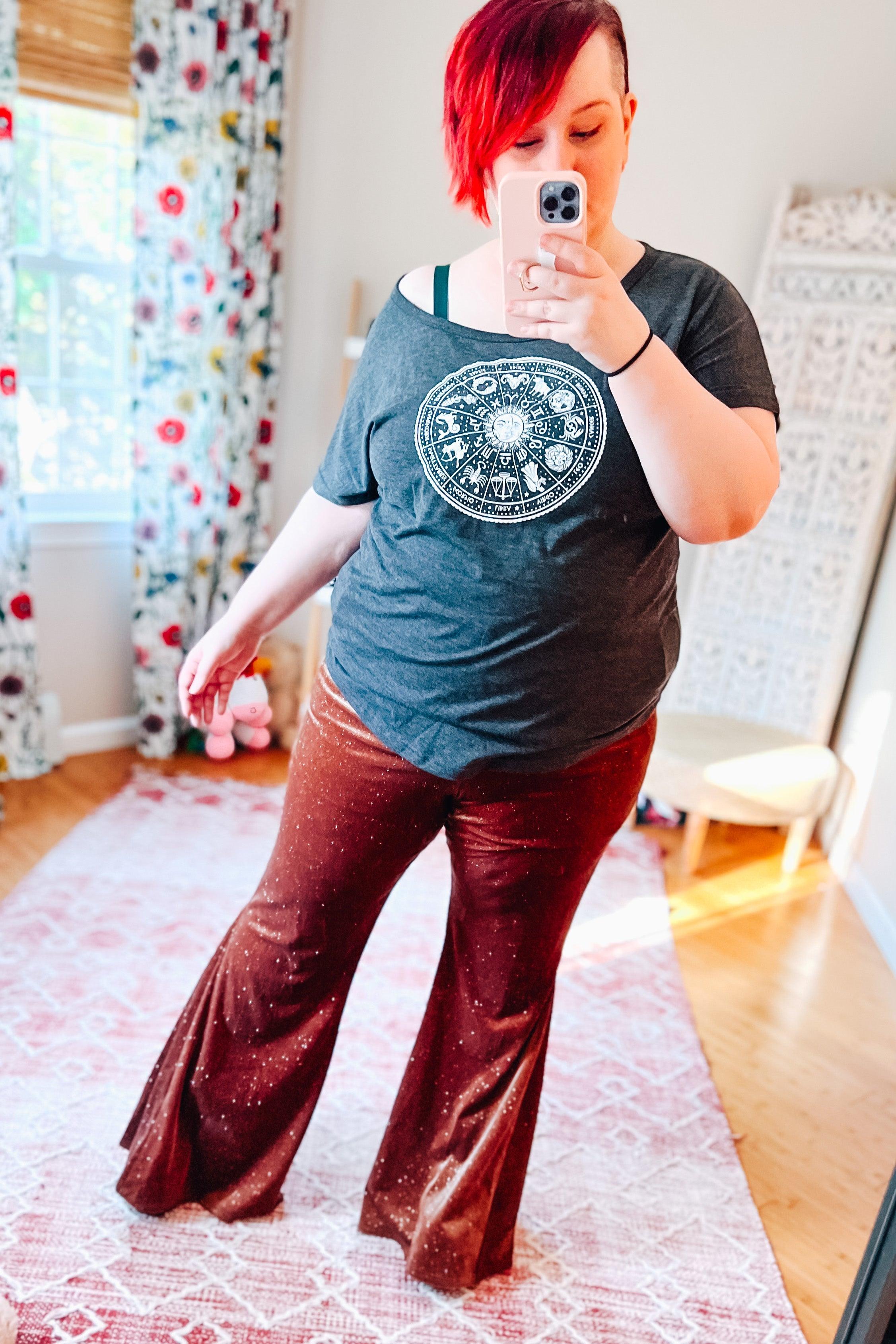 Woman taking mirror selfie wearing rust-colored glitter bell bottoms and grey zodiac print t-shirt indoors