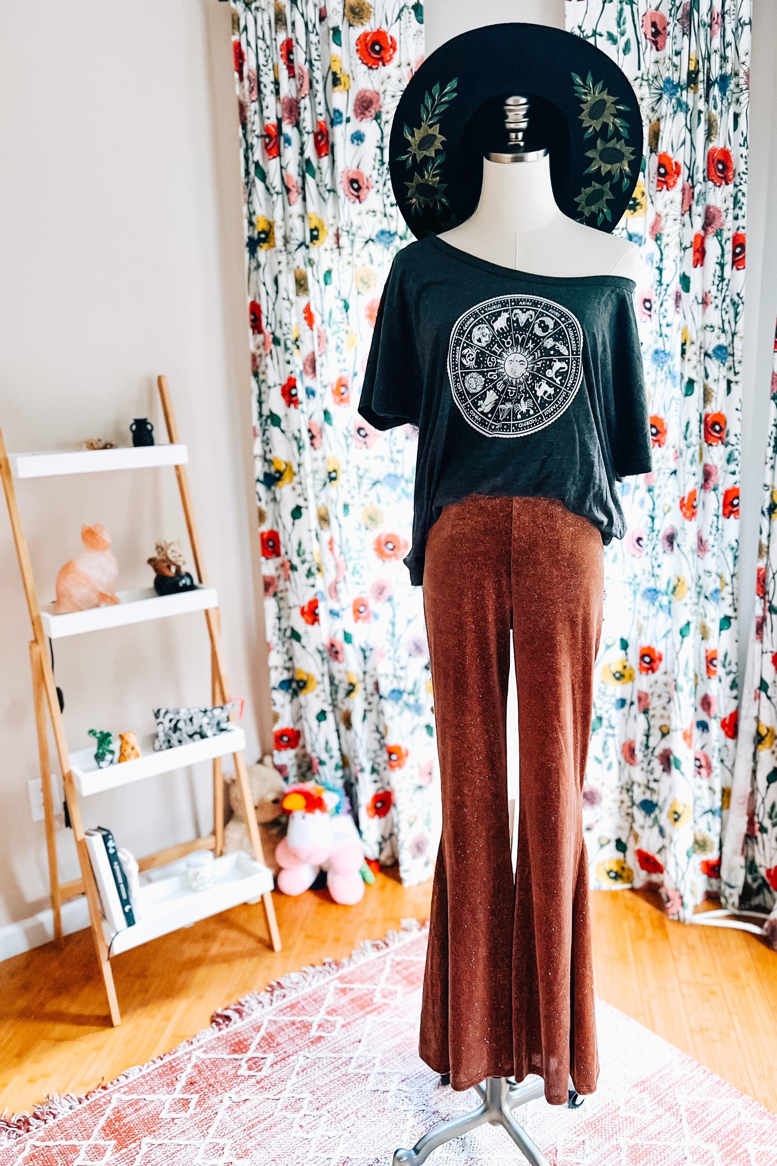 Rust-colored glitter bell bottoms styled with off-shoulder graphic top and wide-brimmed black hat on mannequin indoors