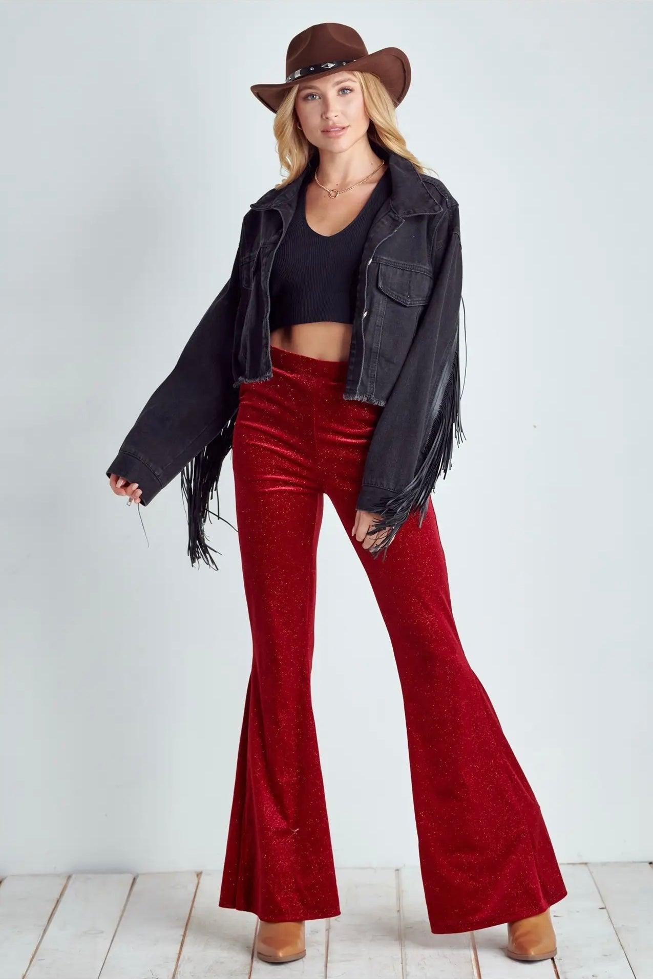 Woman wearing ruby red glitter bell bottoms with cropped black top, fringed black jacket, and brown hat on white floor