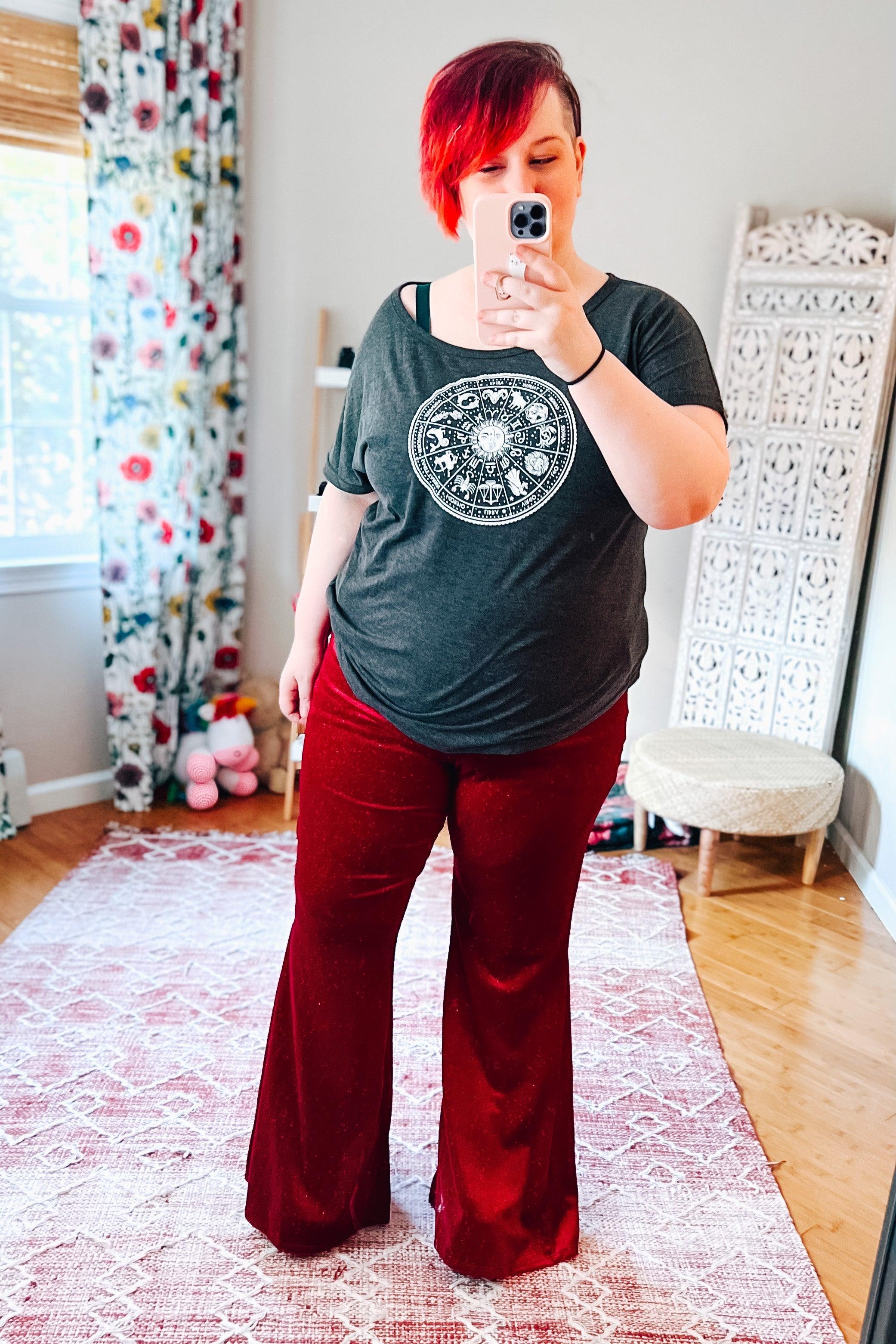 Woman taking selfie wearing ruby glitter velvet bell bottoms and dark gray zodiac graphic t-shirt in cozy colorful room