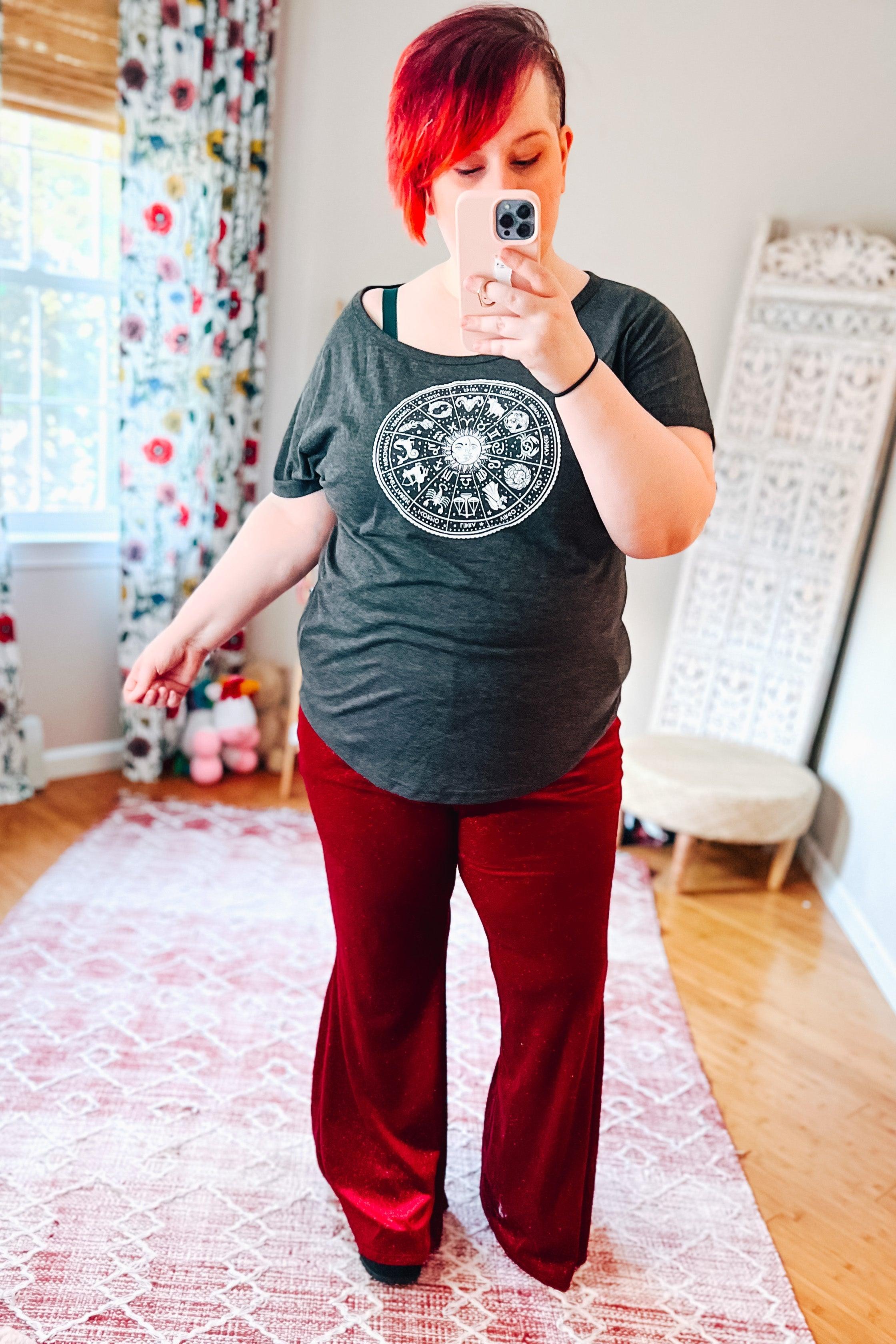 Woman taking mirror selfie wearing ruby red velvet bell bottoms and dark gray zodiac graphic t-shirt in bright room