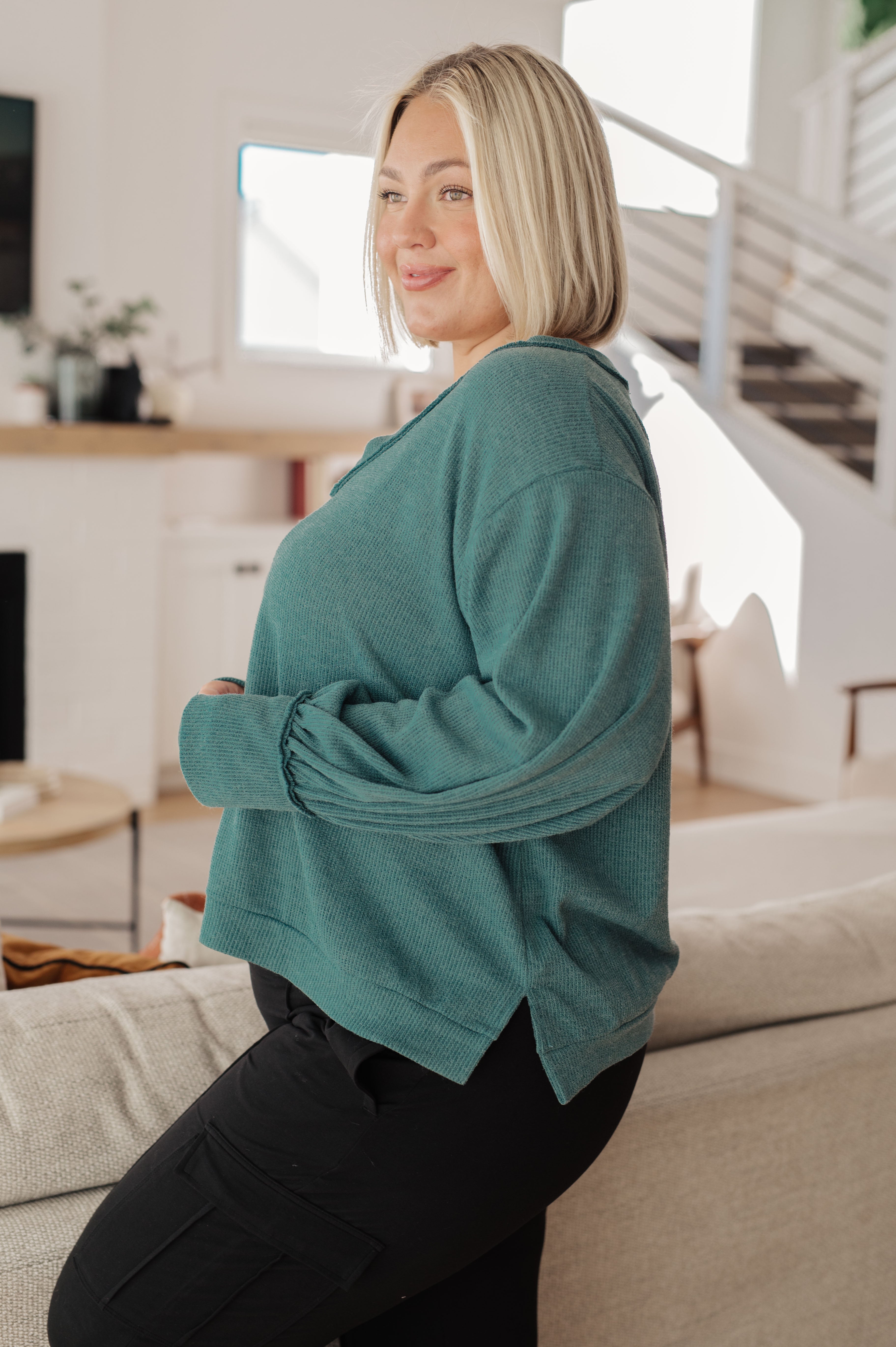 Blonde woman wearing a loose teal knit V-neck top with gathered sleeves sitting on a light gray sofa