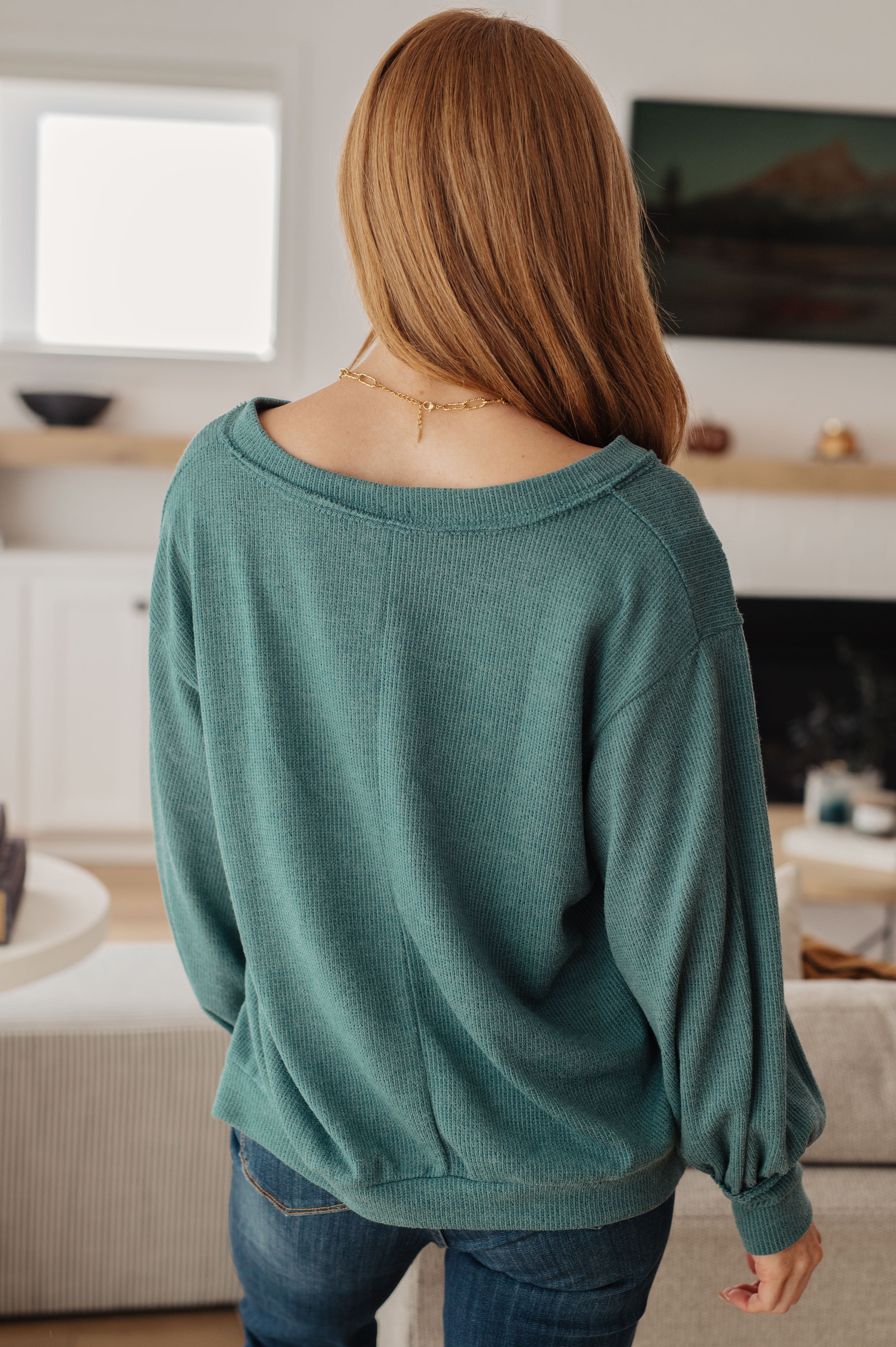 Woman with long red hair wearing teal V-neck top with balloon sleeves in cozy living room setting viewed from back