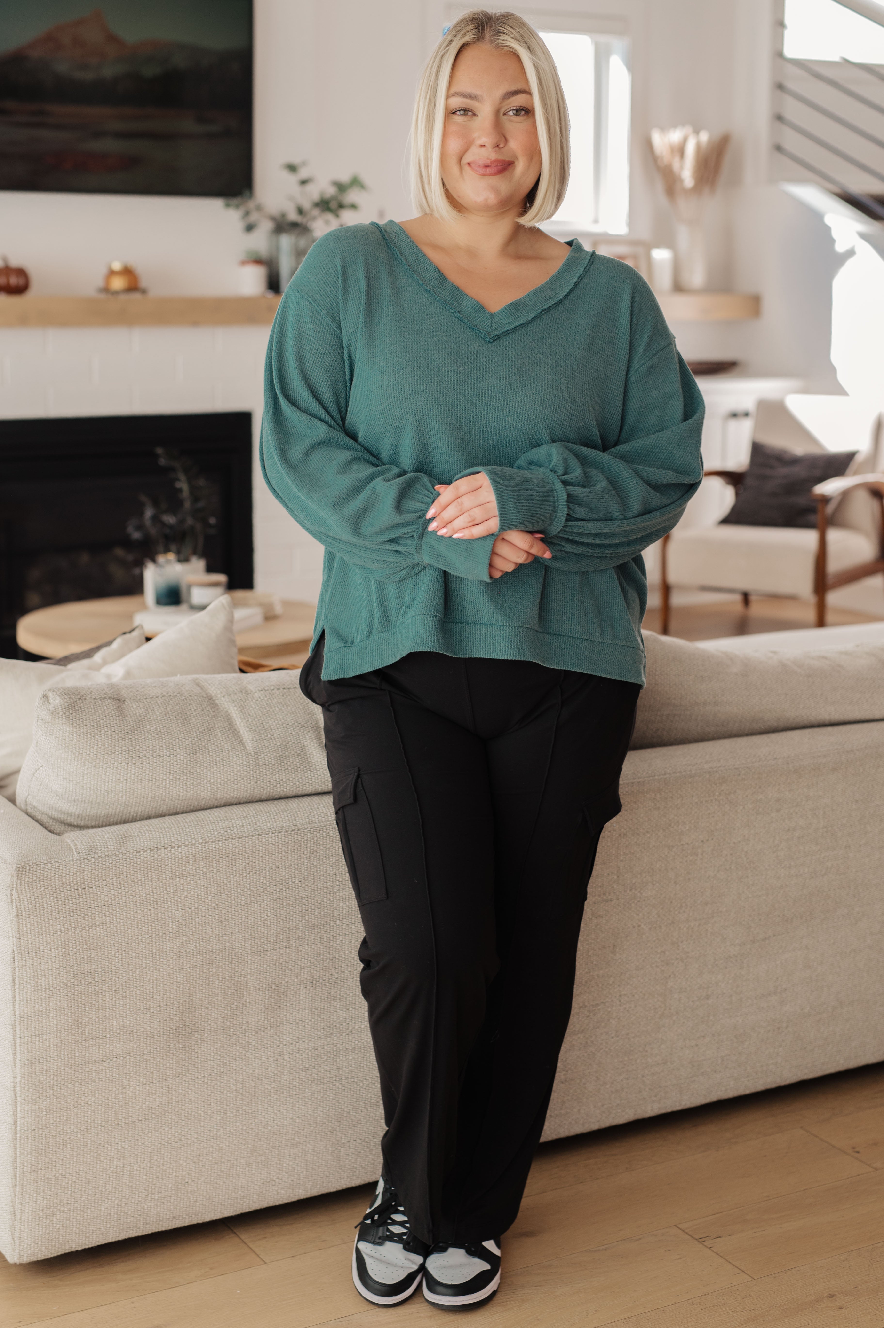 Smiling woman wearing a relaxed teal V-neck sweater with balloon sleeves and black pants in a bright living room
