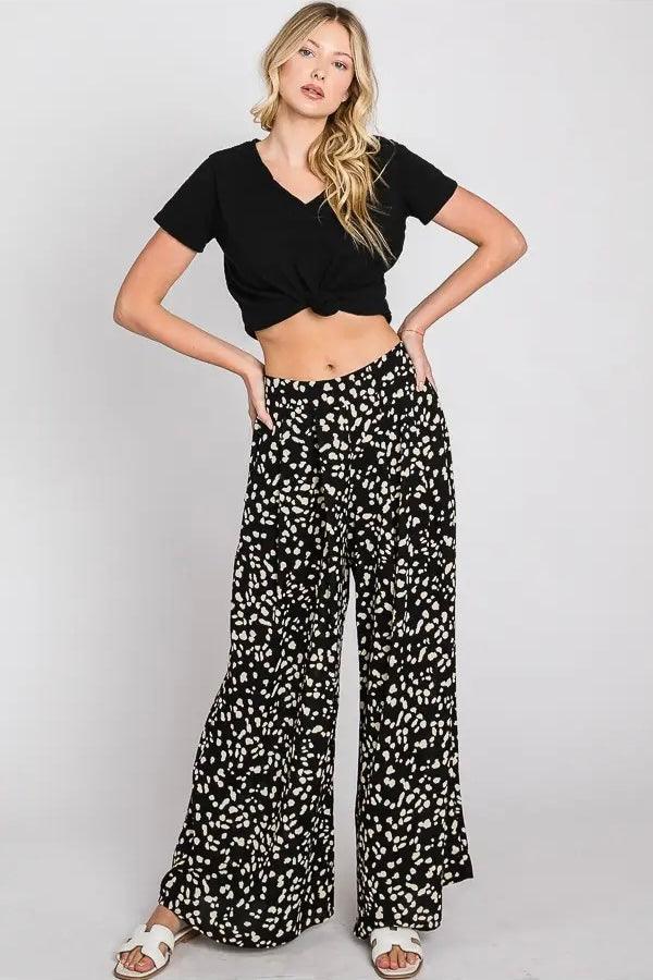 Model wearing black wide leg pants with white abstract dot pattern paired with knotted black crop top and white sandals