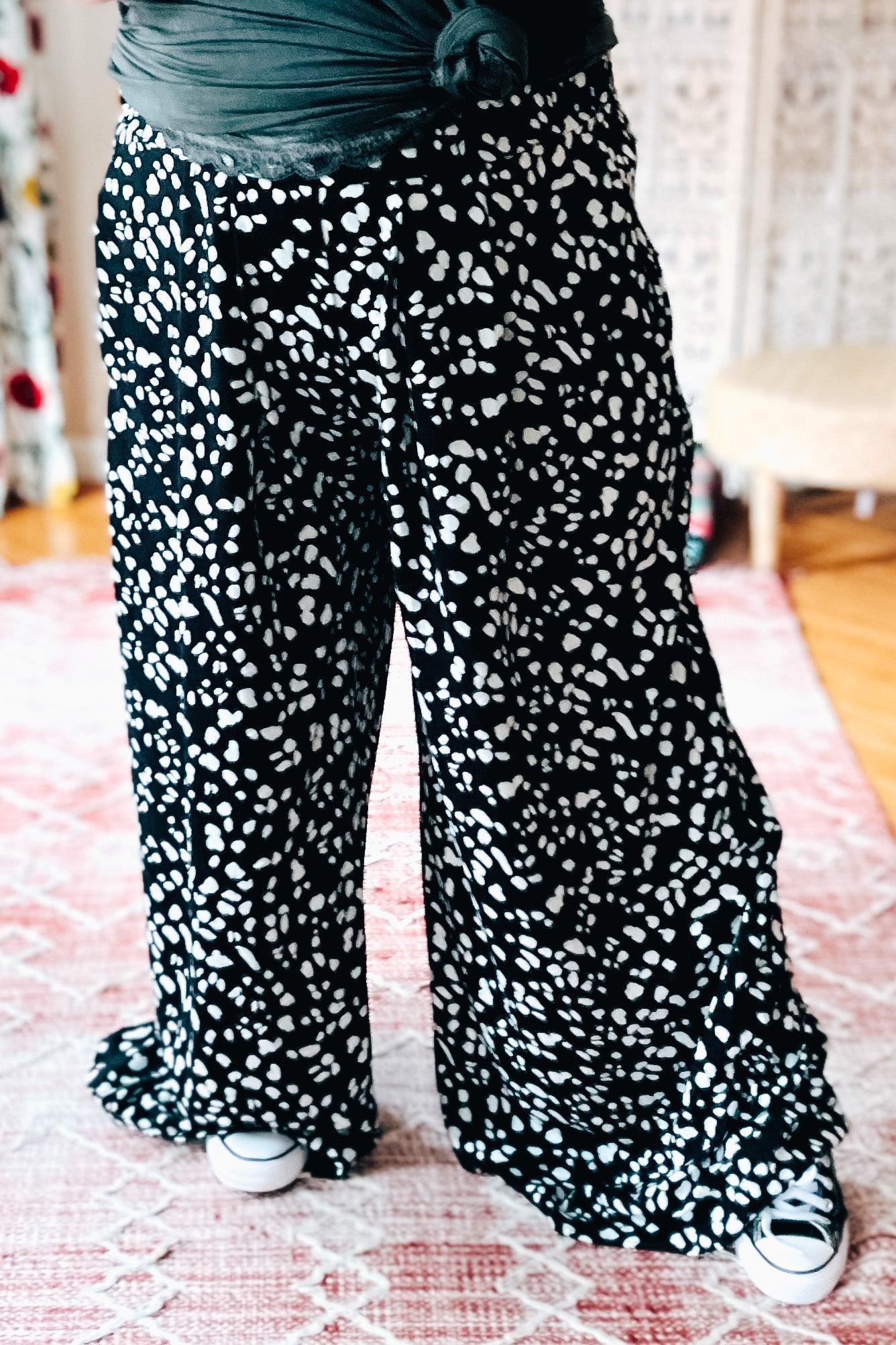 Black wide leg pants with dense white Dalmatian-inspired spots worn with casual sneakers on pink patterned rug