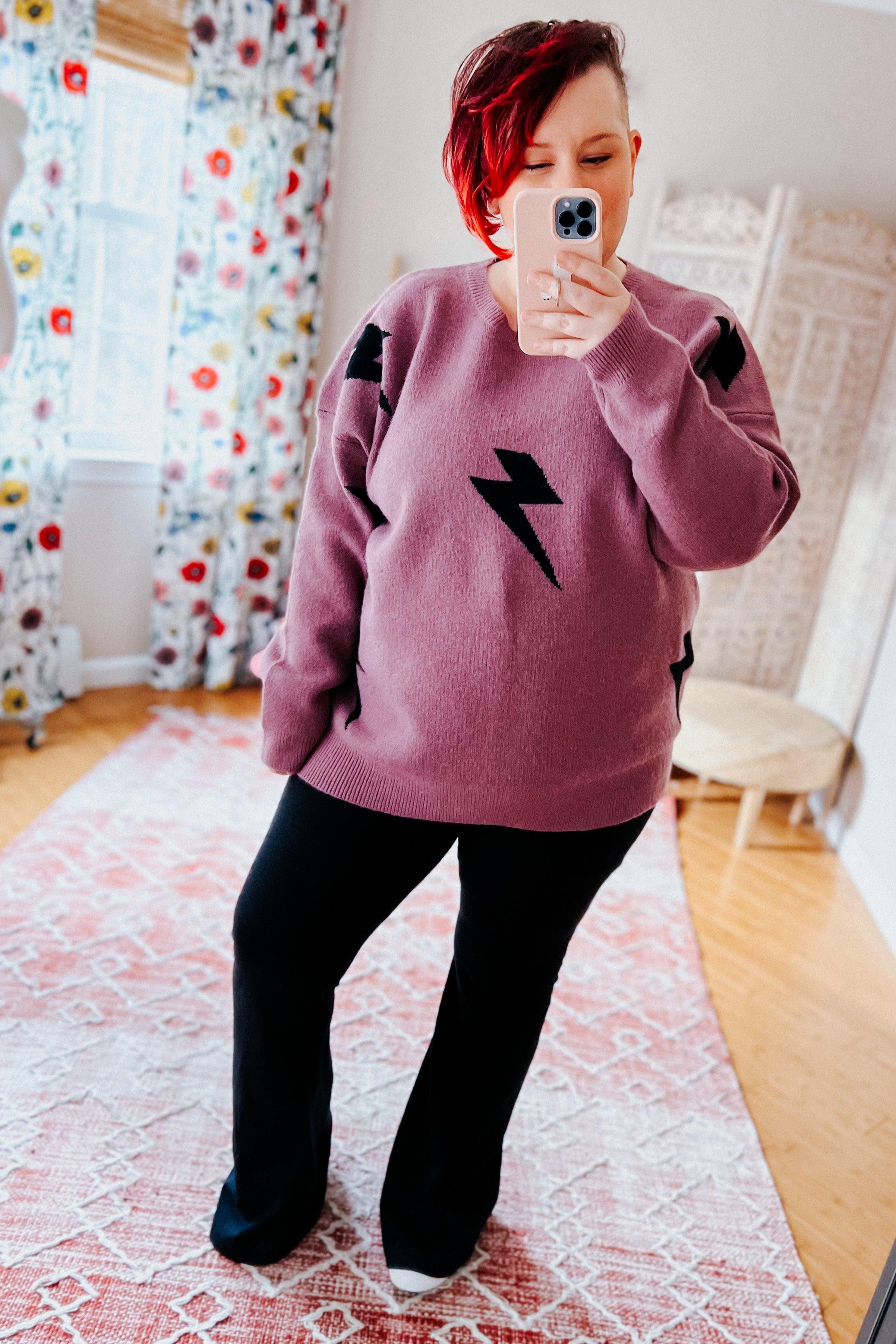 Woman wearing a purple sweater with black lightning bolt designs and black flared leggings, standing in a room with floral curtains.