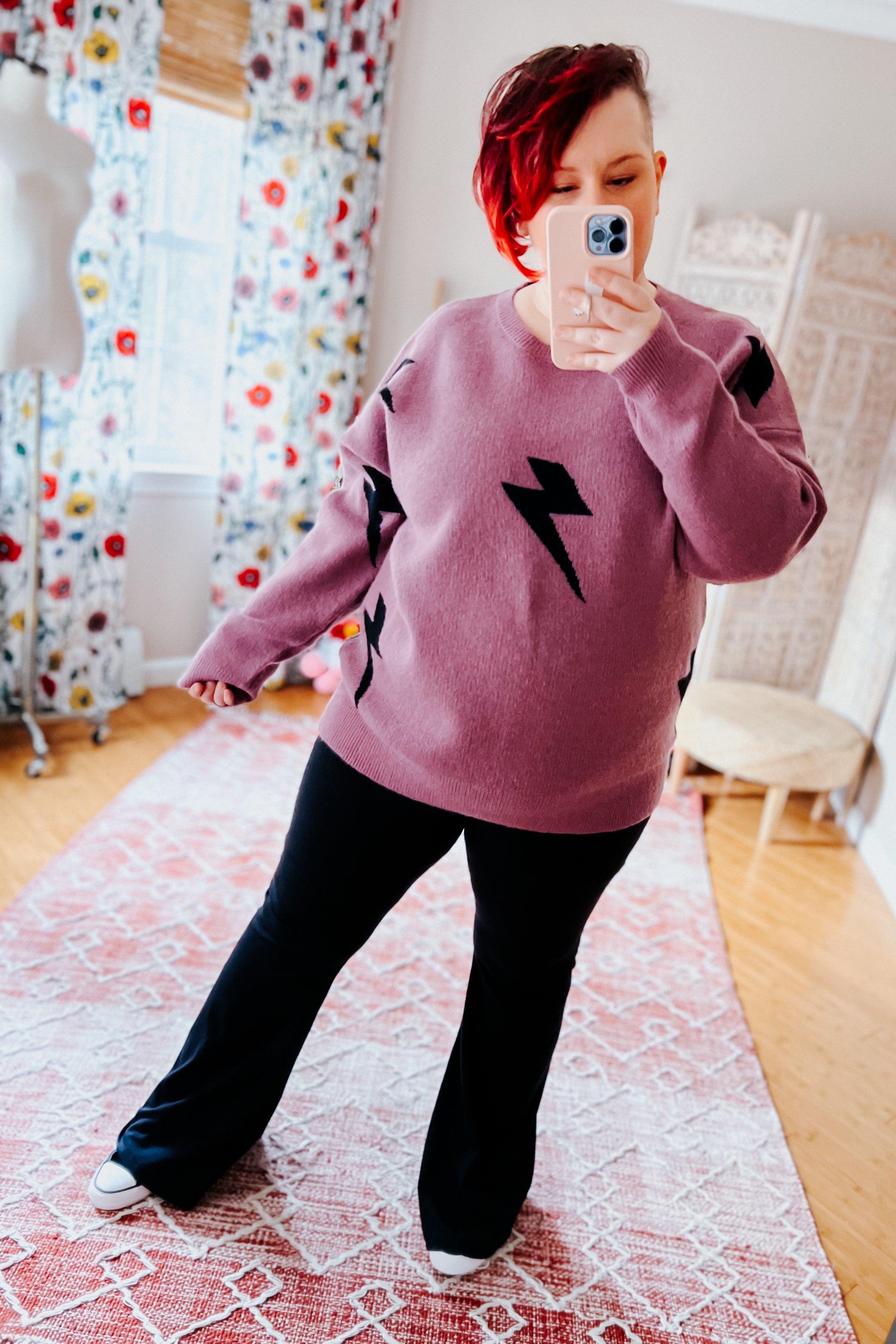 Woman wearing a purple sweater with black lightning bolt designs and black flared leggings, standing in a room with floral curtains.