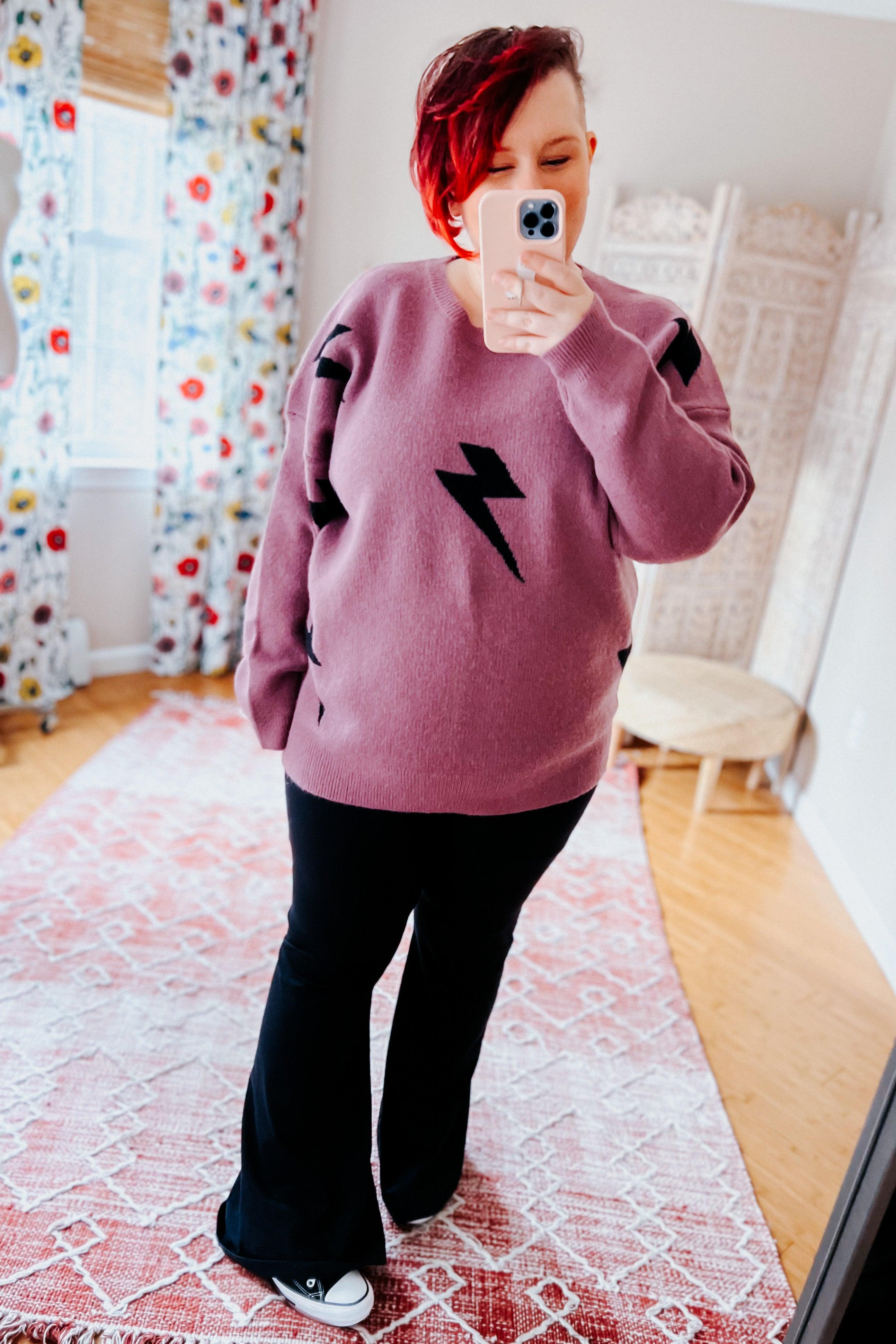 Woman wearing a purple sweater with black lightning bolt designs and black flared leggings, standing in a room with floral curtains.