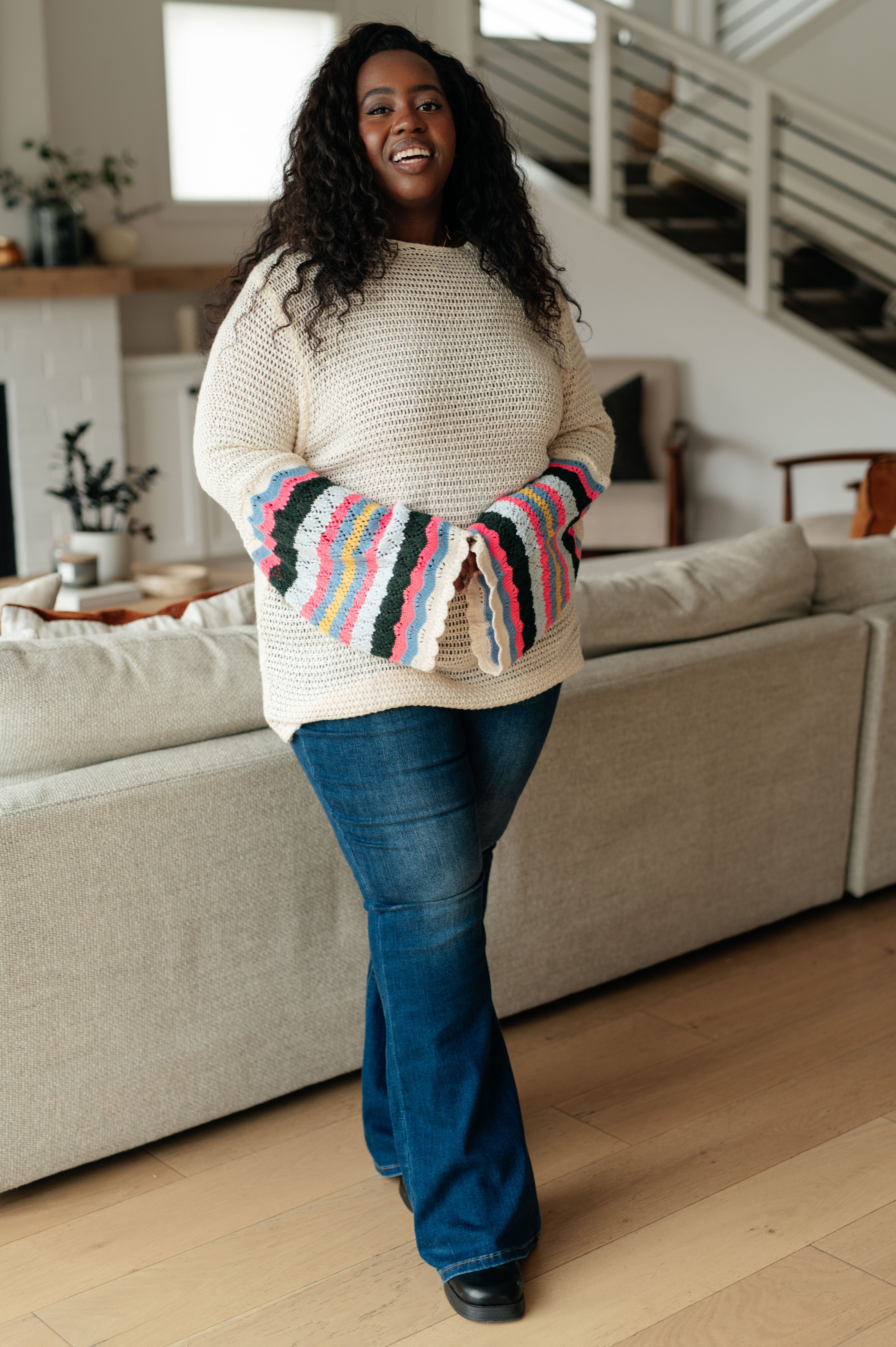 Smiling woman indoors wearing cream knit sweater with colorful striped bell sleeves and dark blue jeans standing by sofa