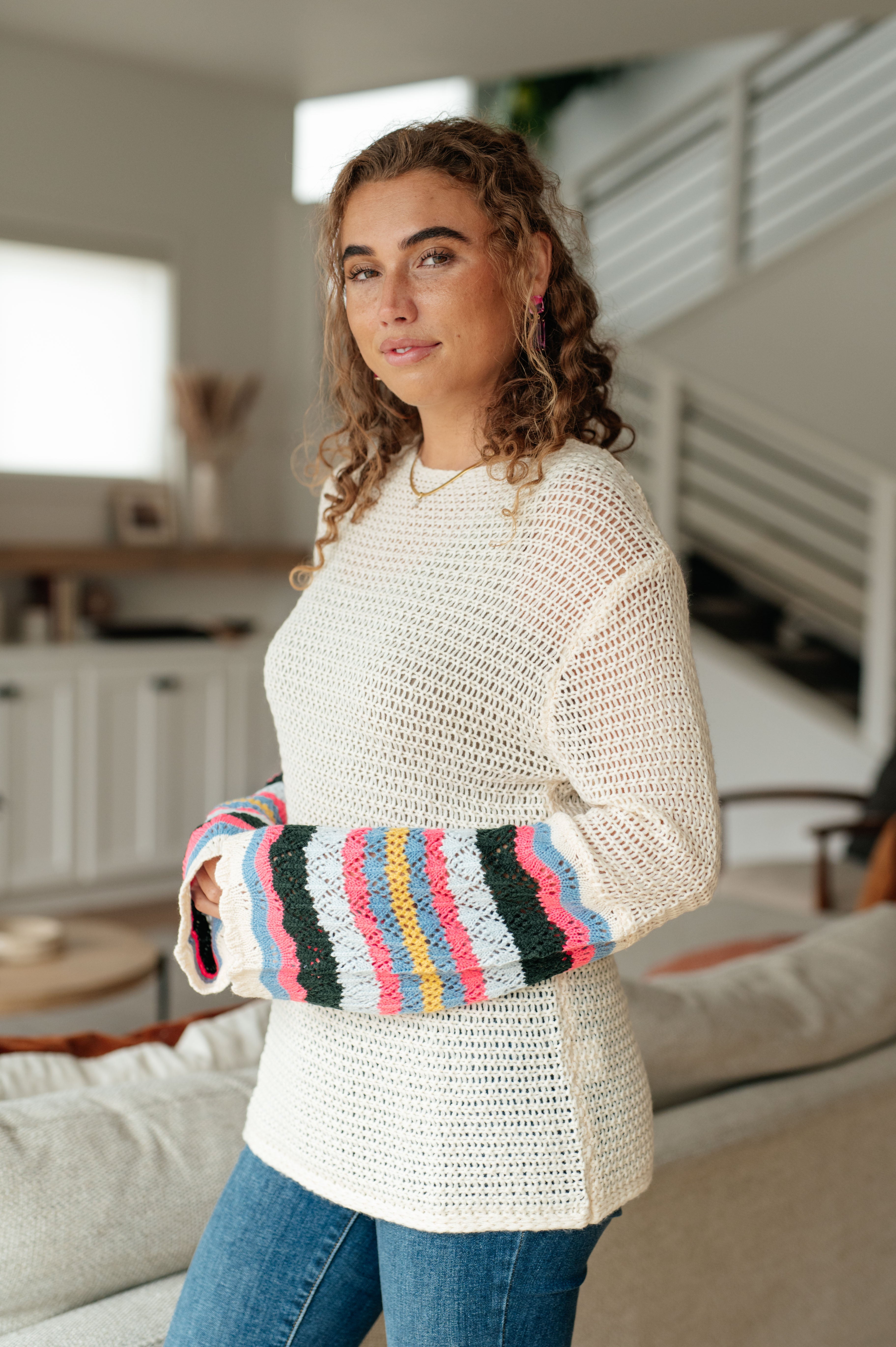 Woman indoors wearing cream crocheted sweater with multicolored striped sleeves and blue jeans standing by sofa