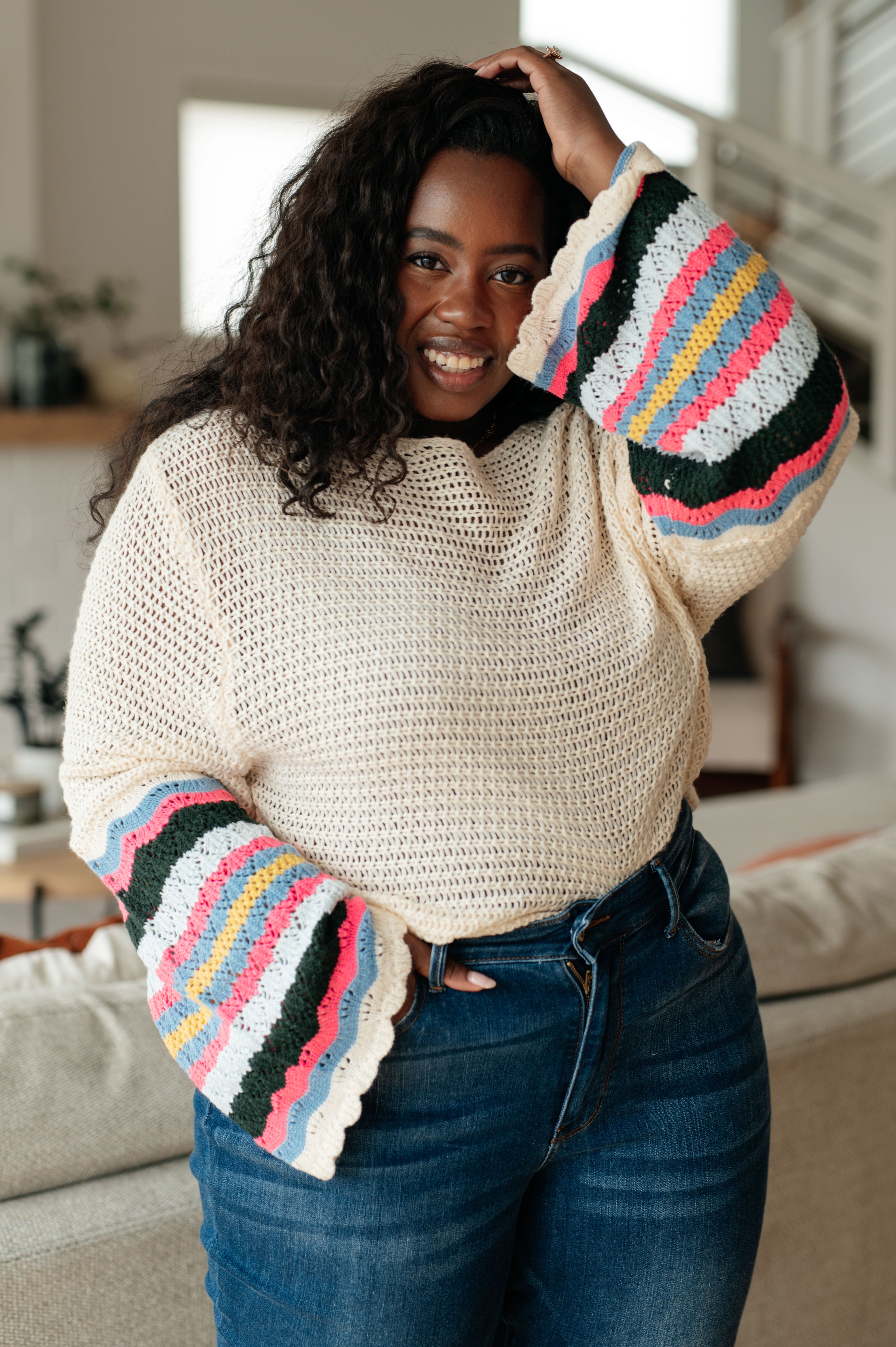Smiling woman wearing cream crochet sweater with colorful striped bell sleeves and high-waisted dark blue jeans indoors