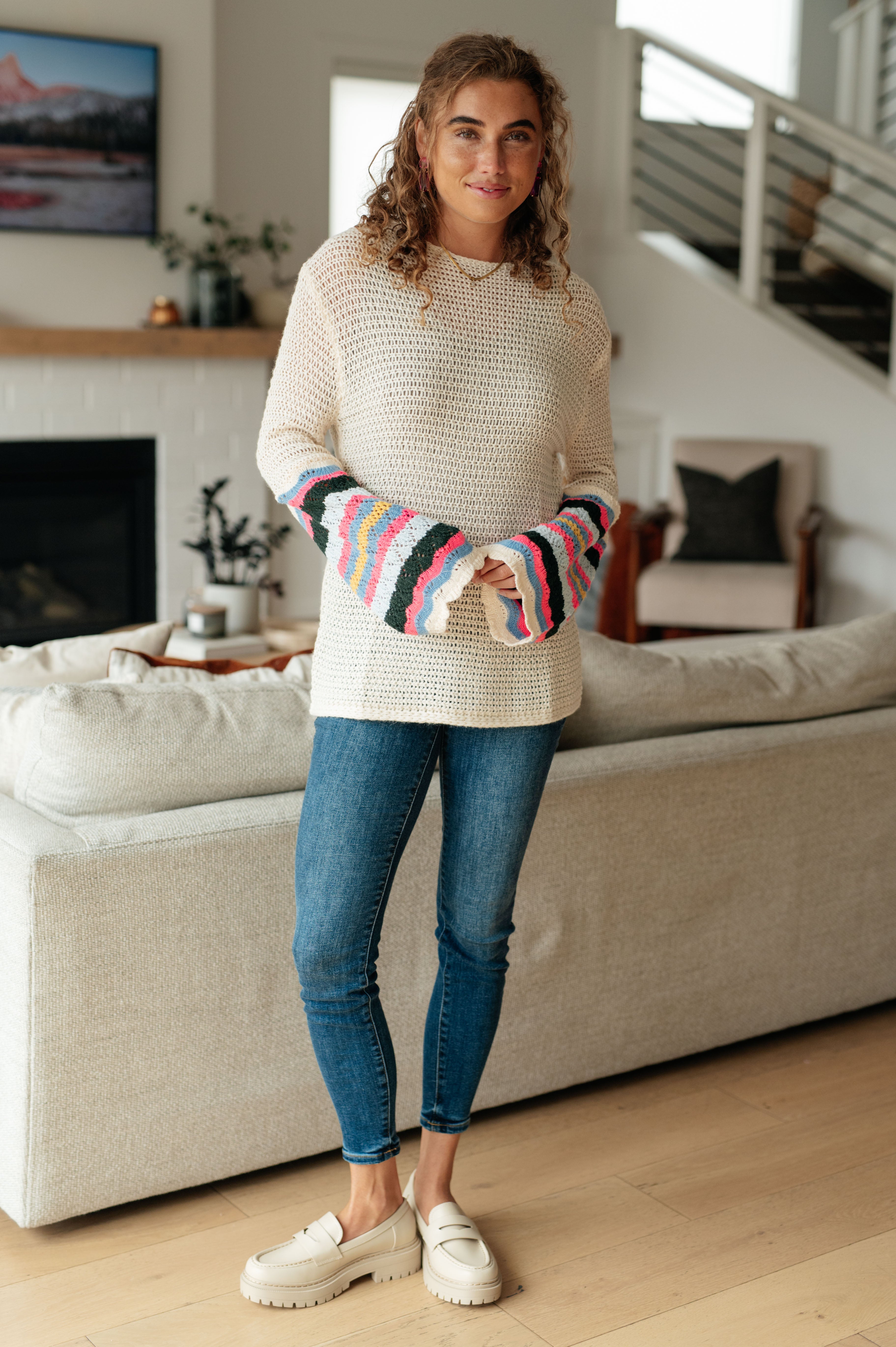 Woman standing indoors wearing cream knit sweater with colorful striped bell sleeves and skinny blue jeans