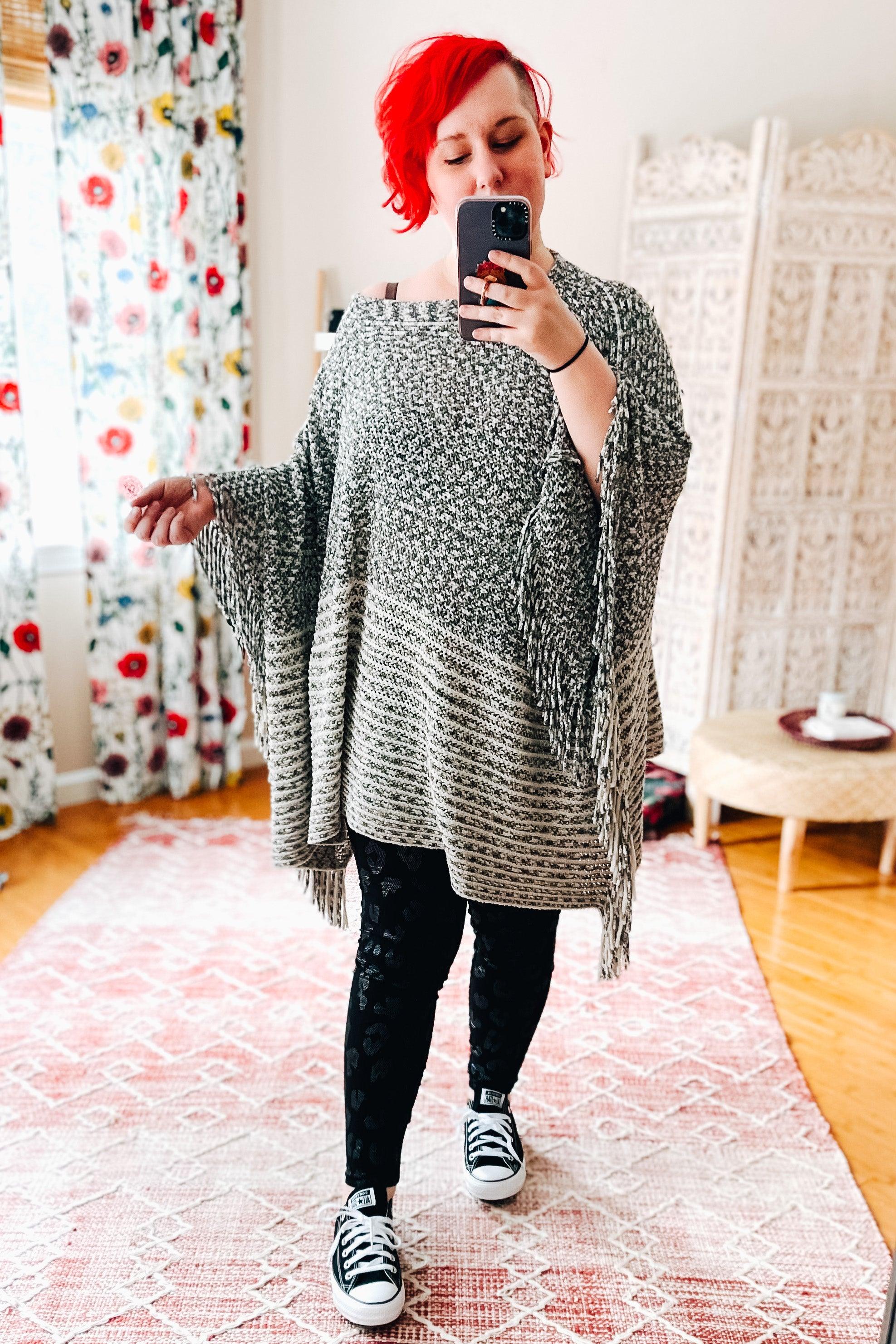 Woman with bright red hair wearing a gray knit poncho and black leopard foil print leggings taking a selfie indoors