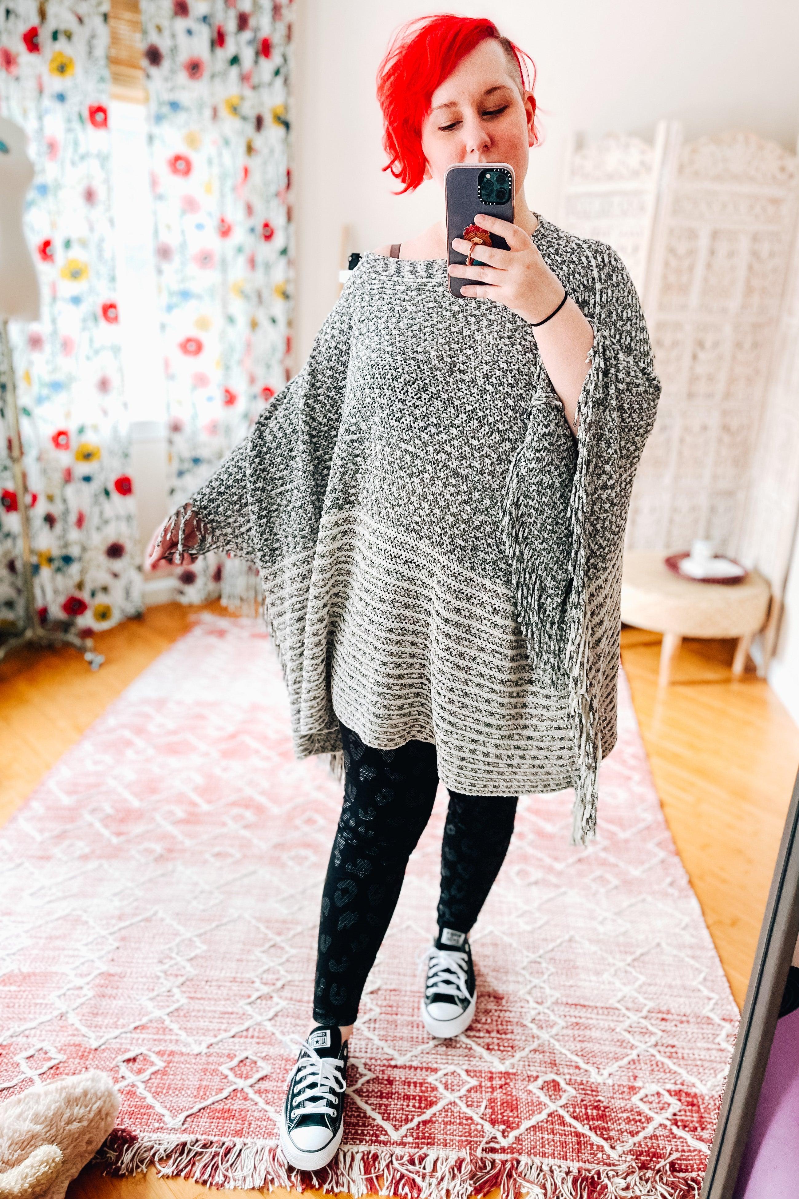Woman with red hair wearing a gray woven poncho and black leopard print leggings taking a mirror selfie indoors