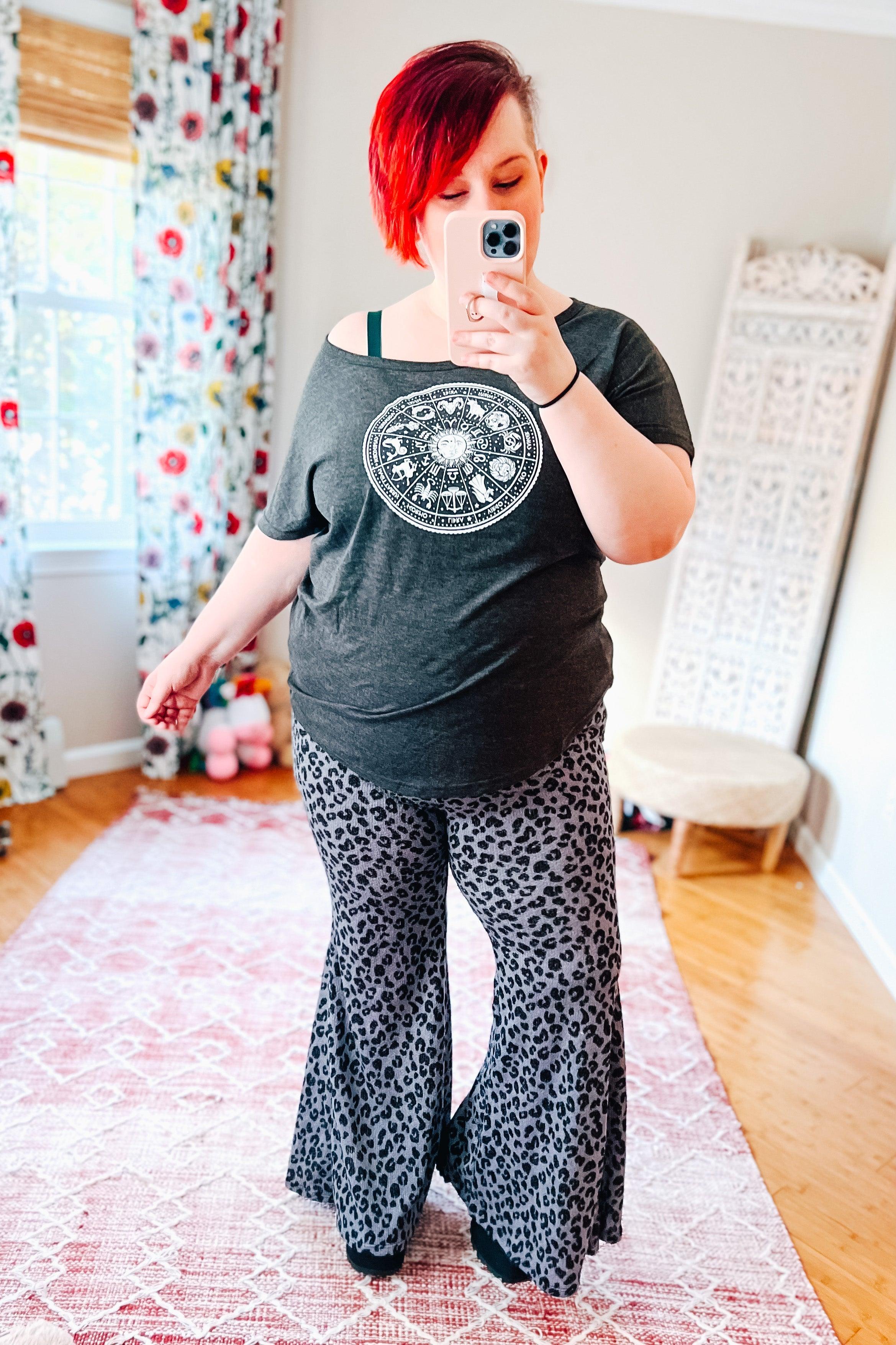 Woman taking selfie wearing wide-legged leopard print pants and off-shoulder dark gray graphic top indoors