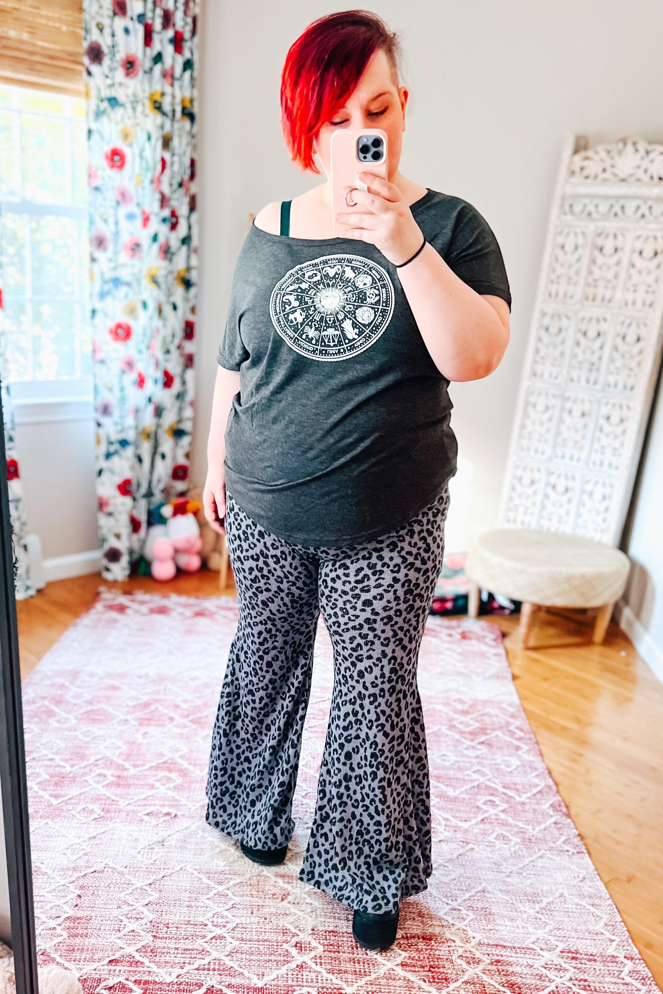 Woman taking mirror selfie wearing black and blue leopard print flared bell bottom pants and dark graphic t-shirt indoors