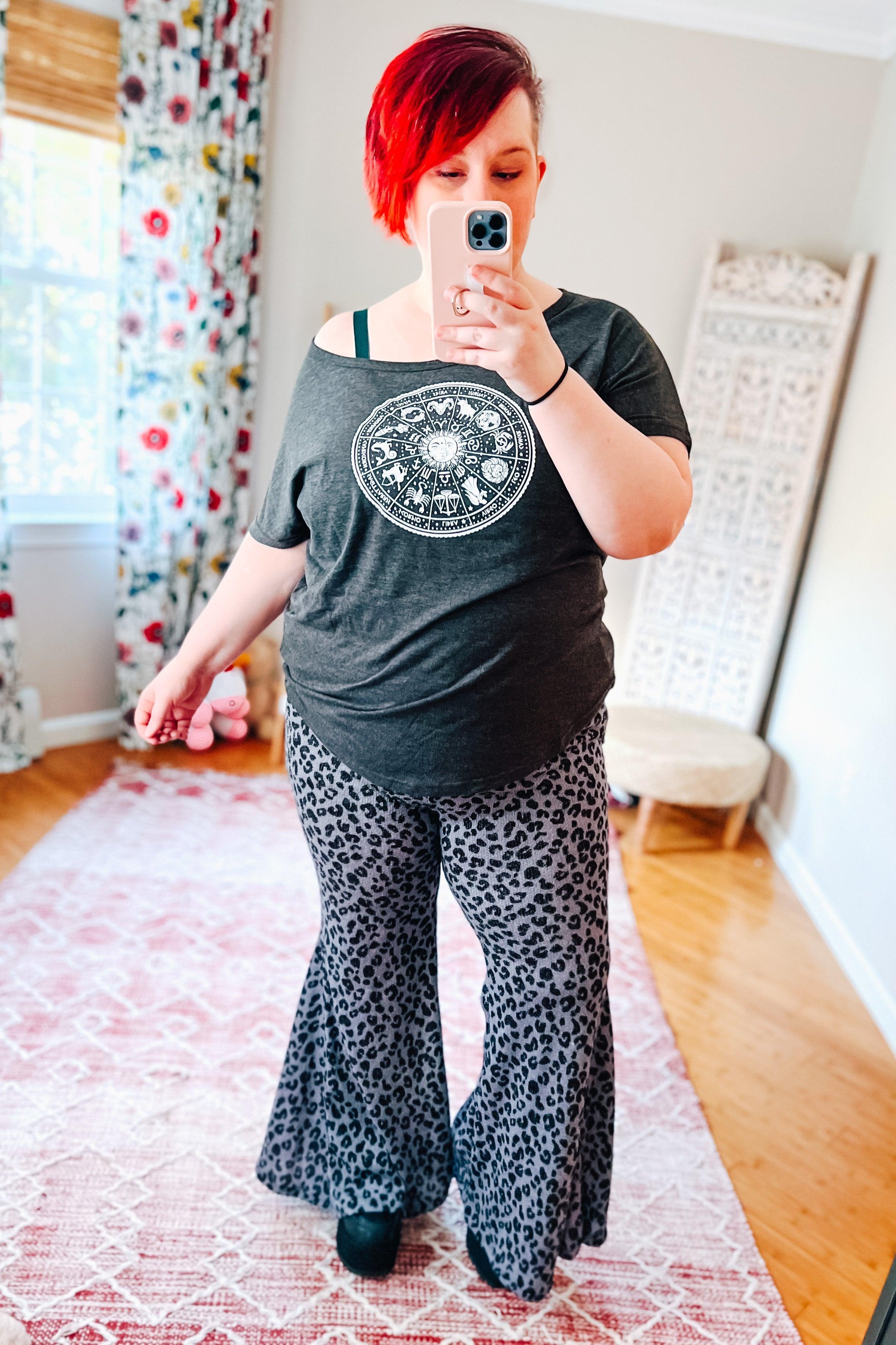 Woman taking mirror selfie wearing black and blue leopard print bell bottom pants and dark gray graphic off-shoulder top indoors