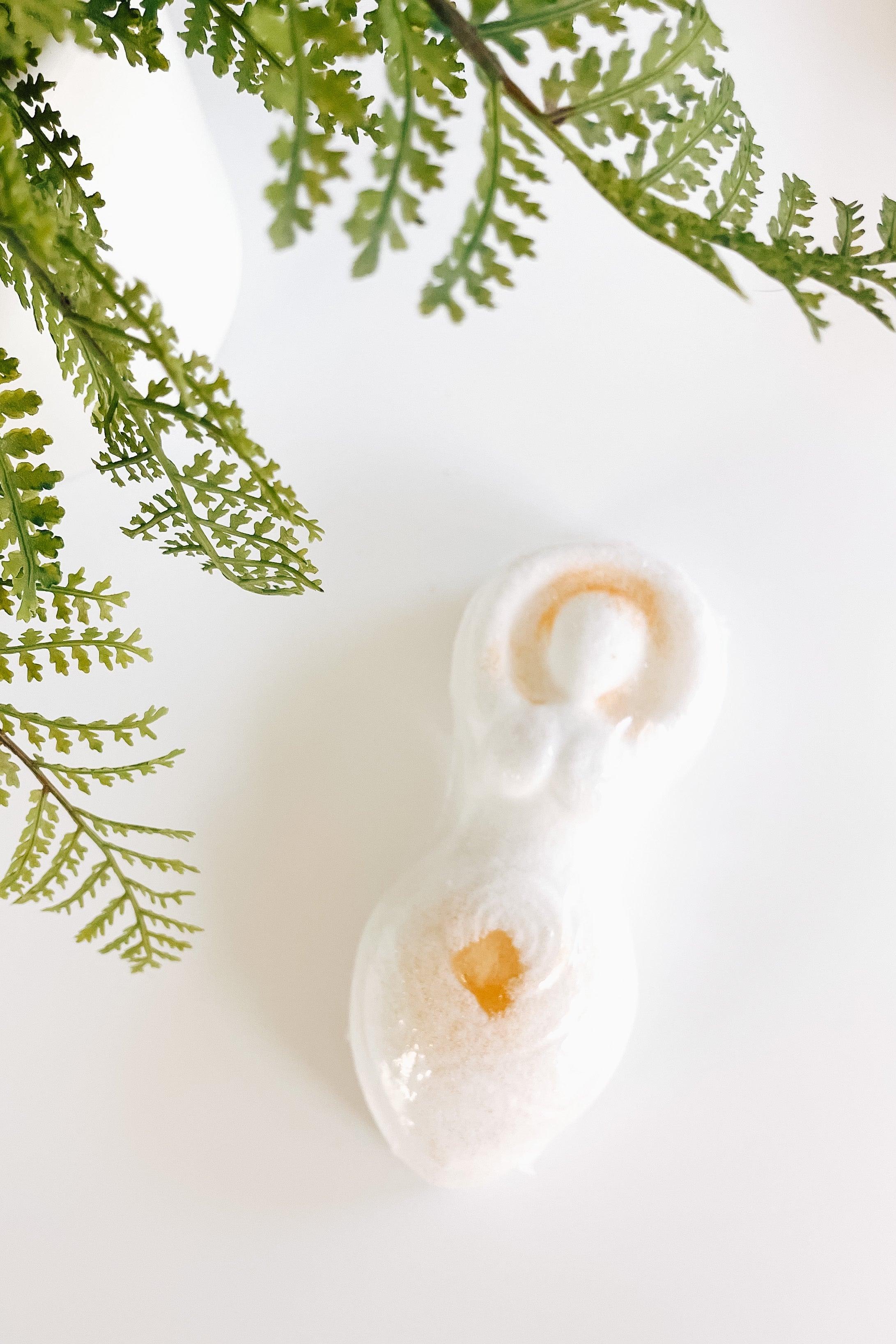 Cream goddess-shaped bath bomb with warm citrine-colored crystal details surrounded by green fern leaves on white background
