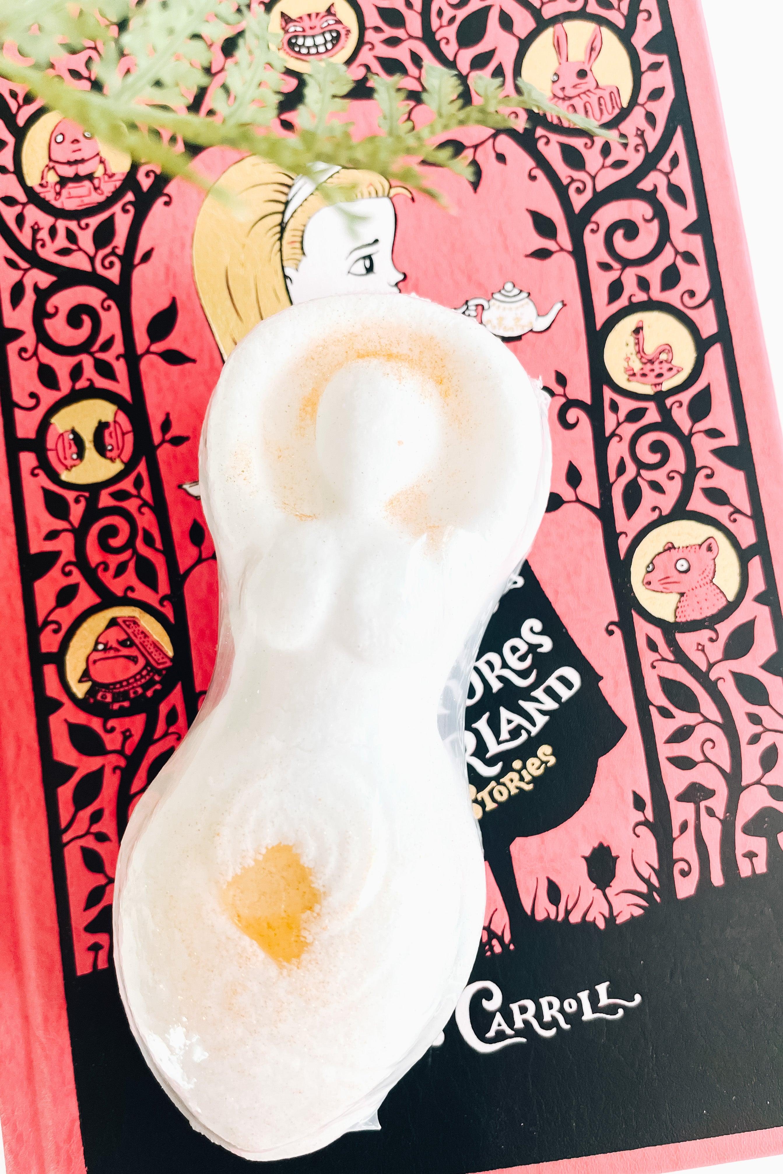 Cream goddess-shaped bath bomb with subtle golden highlights resting on colorful illustrated book beneath green fern leaves