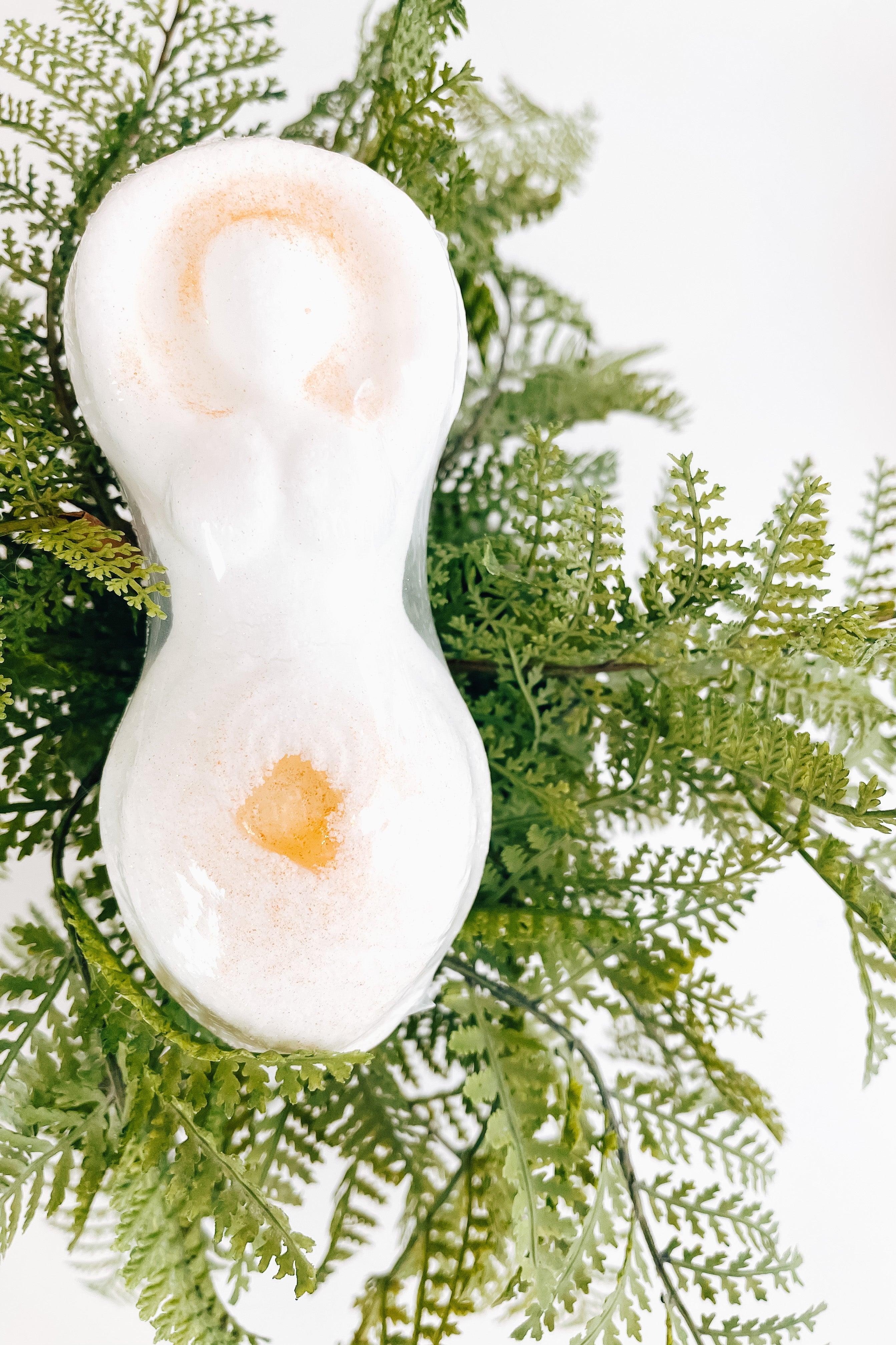 Cream bath bomb with golden citrine crystal detail resting atop lush green fern leaves on white surface