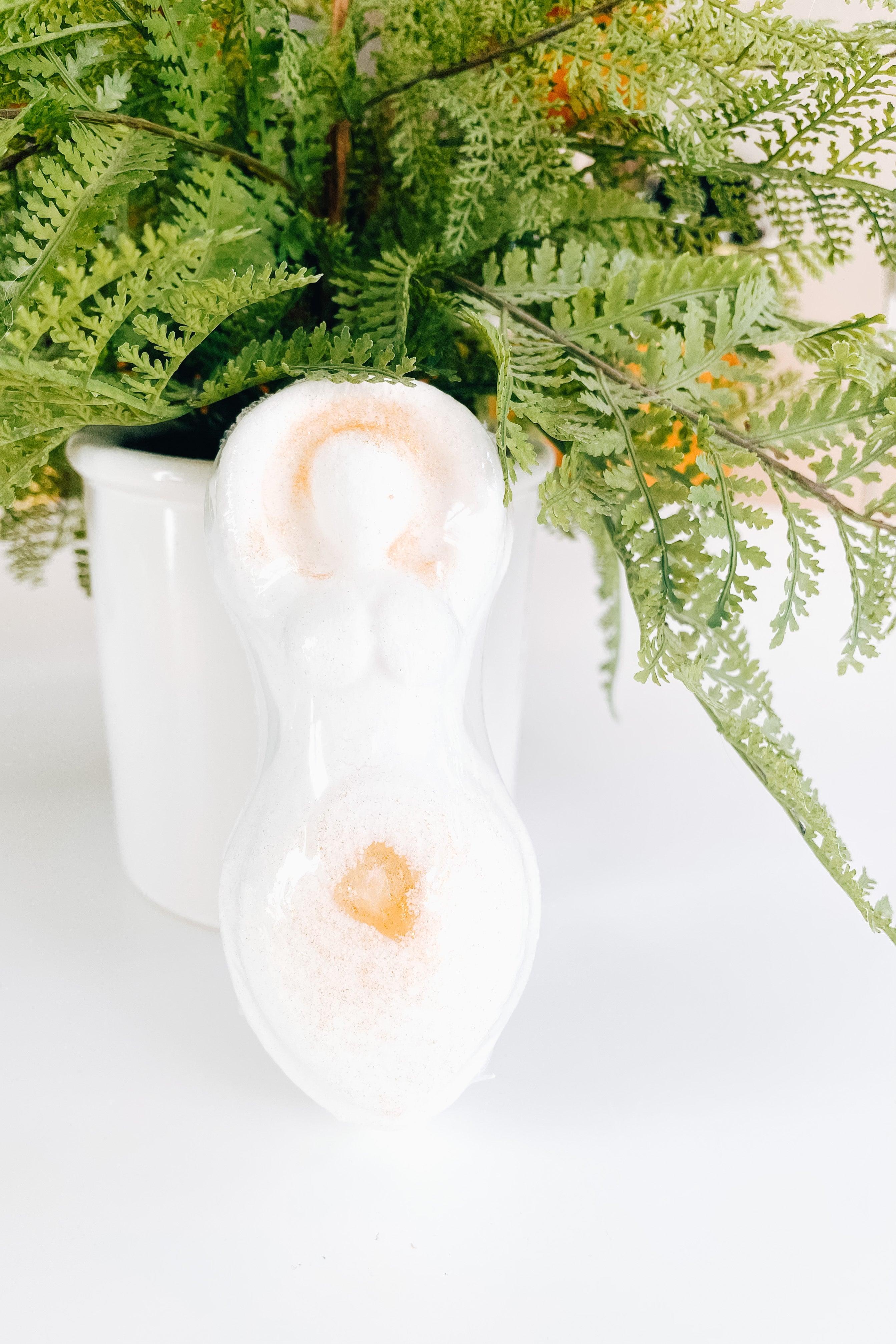 Cream colored bath bomb shaped like a goddess figurine with citrine crystal detail resting against a white pot with green fern leaves