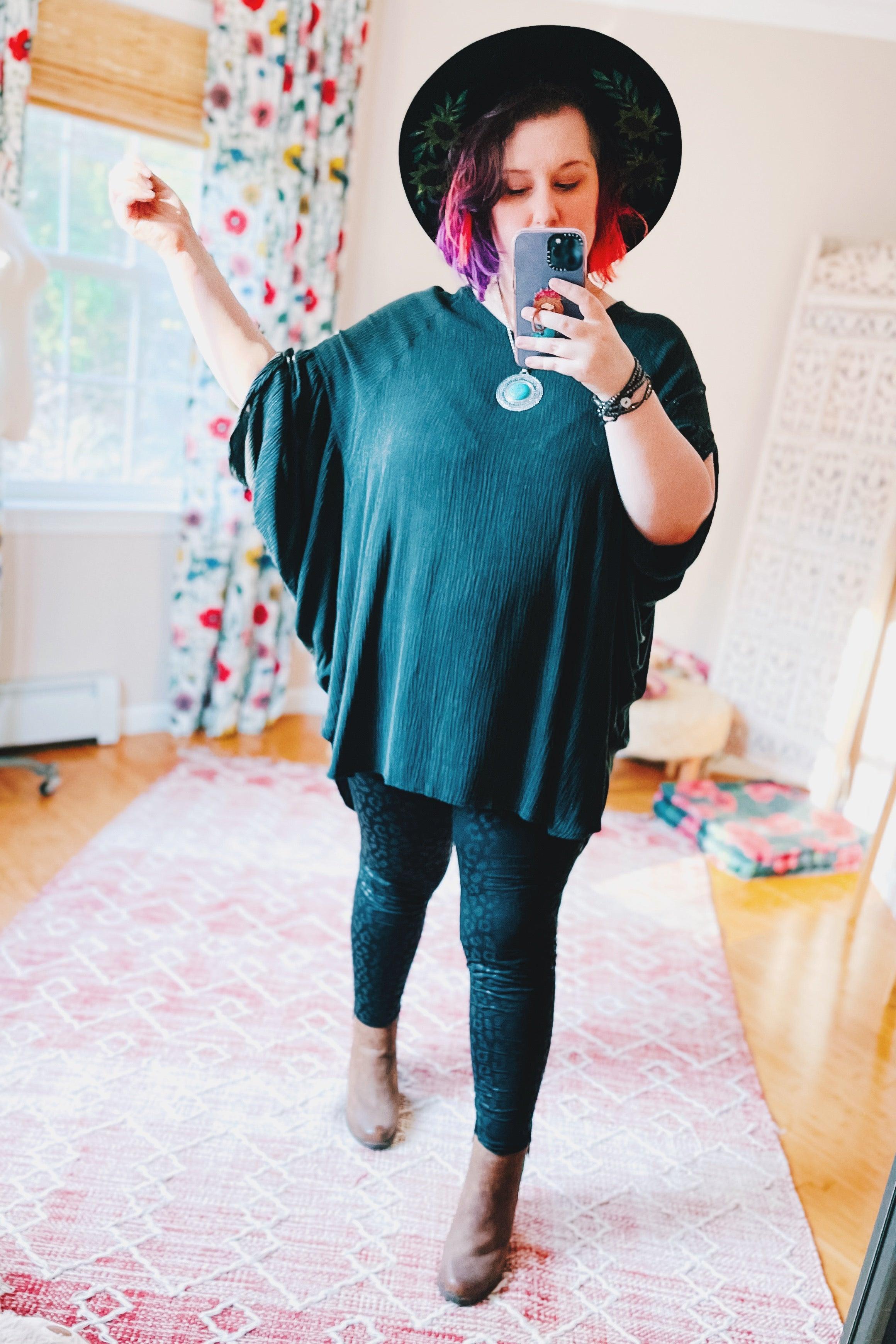 Woman taking mirror selfie wearing charcoal loose tunic with wide sleeves black hat and patterned black leggings indoors