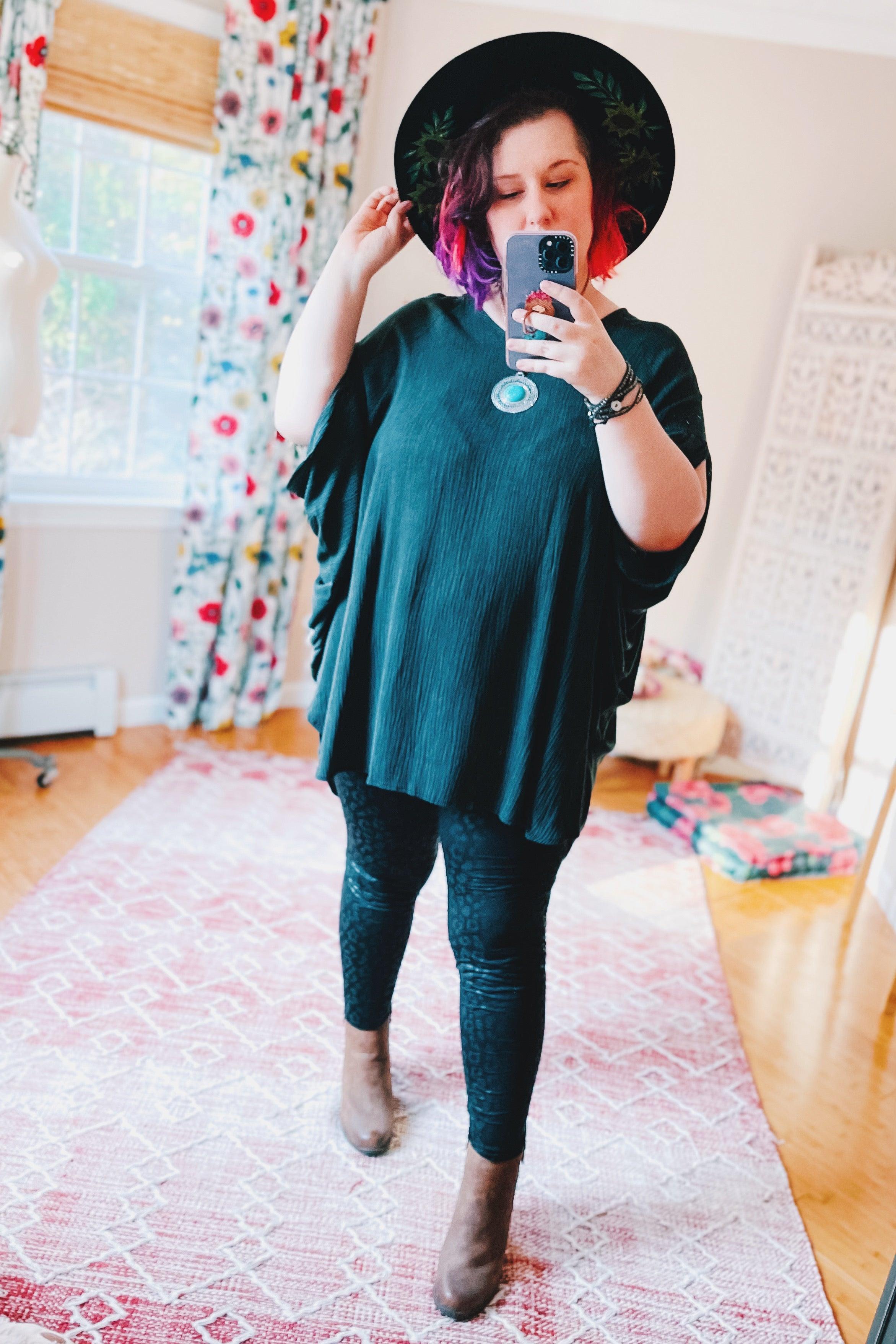 Woman taking mirror selfie wearing loose charcoal tunic with wide sleeves, black hat, patterned leggings, and brown ankle boots indoors
