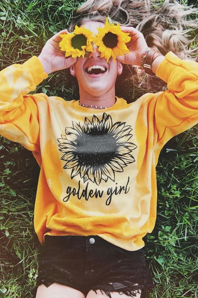 Woman lying on grass wearing yellow bleached pullover with large sunflower graphic and golden girl lettering holding sunflowers over eyes