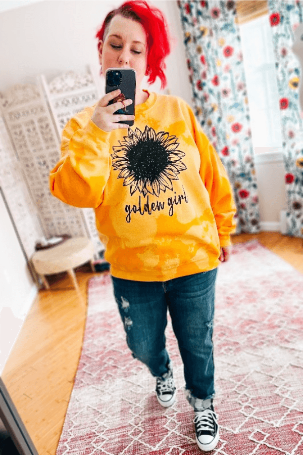 Woman with red hair wearing a yellow pullover with black sunflower graphic and golden girl text taking a mirror selfie indoors
