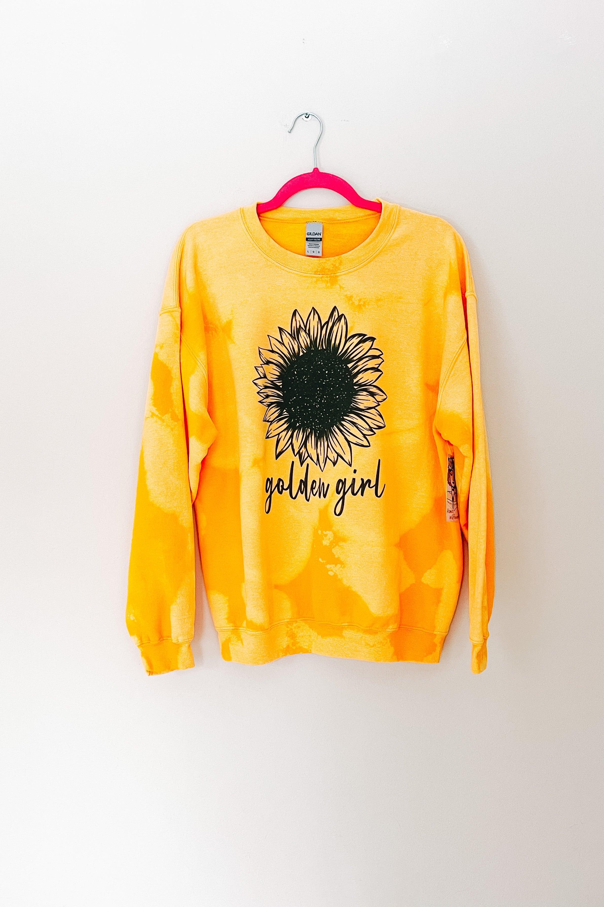Yellow bleached pullover with a large black sunflower graphic and golden girl text hanging on a pink hanger against a white wall